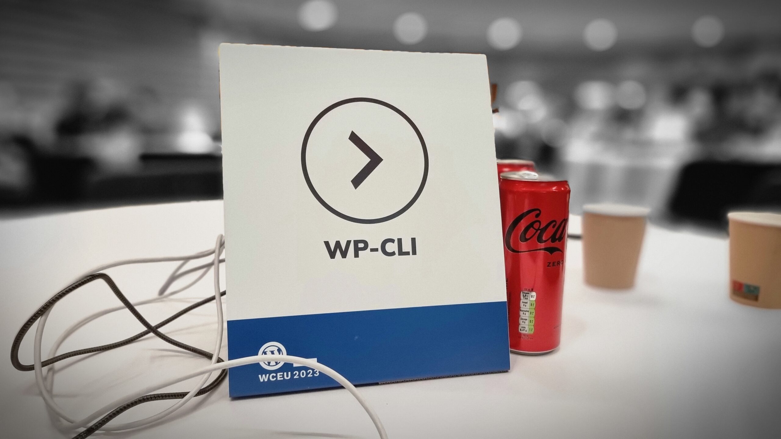 Full image of A table placeholder labelled "WP-CLI". It and s handful of Coke cans and disposable coffee cups are picked-out in colour on an otherwise monochrome and blurred picture.
