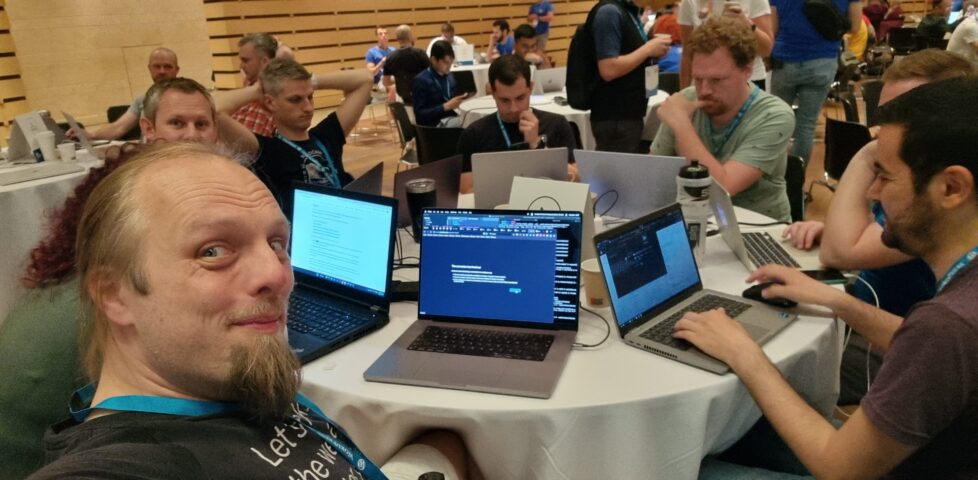 Dan takes a selfie from a round table covered in laptops, with people hacking at them.