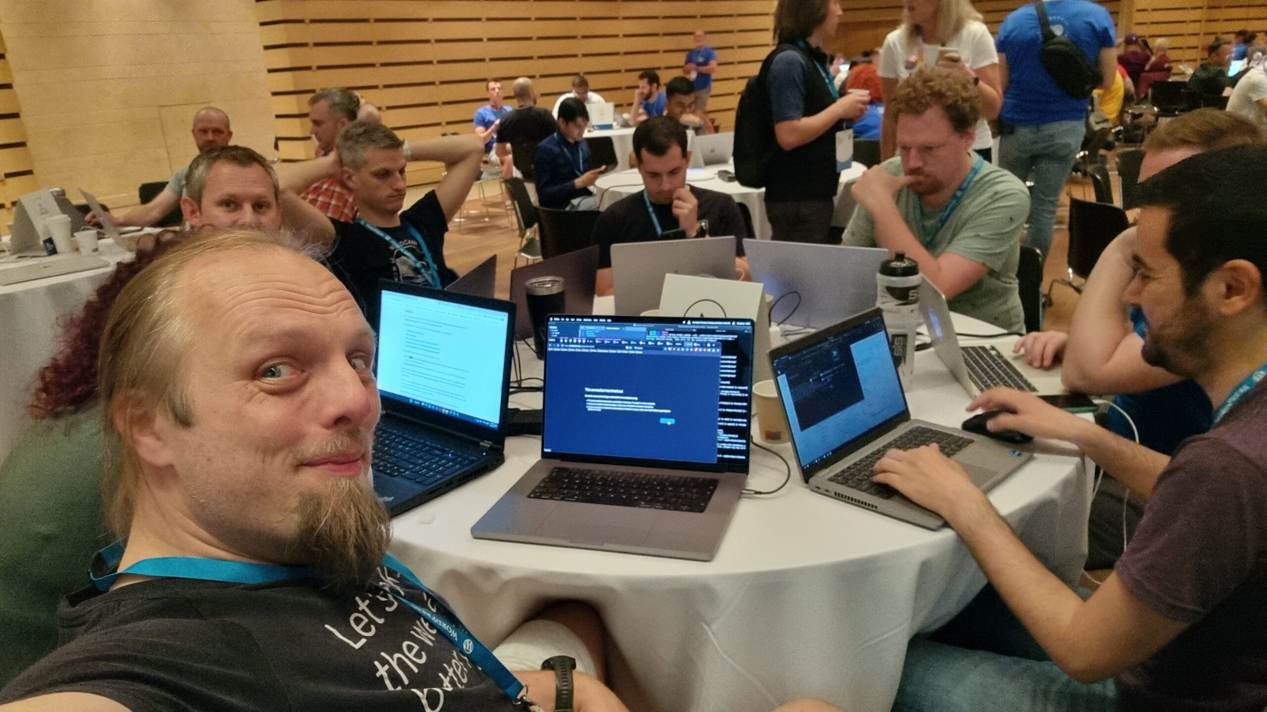 Full image of Dan takes a selfie from a round table covered in laptops, with people hacking at them.