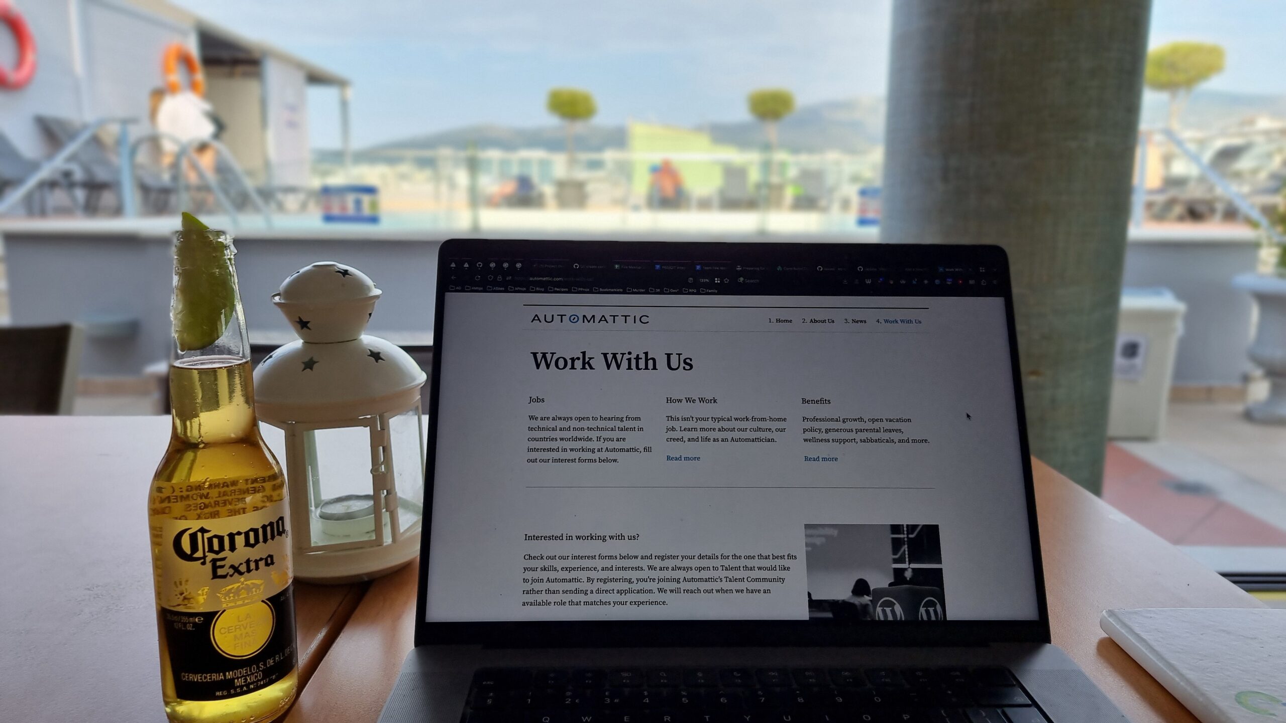 Full image of MacBook showing an Automattic "Work For Us" web page, alongside a bottle of Corona Extra. A rooftop terrace garden and swimming pool can be seen in the background.