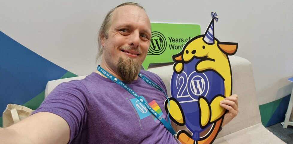 Dan, smiling, wearing a purple t-shirt with a WordPress logo and a Pride flag, hugs a cut-out of a Wappu (itself hugging a "WP 20" balloon and wearing a party hat).