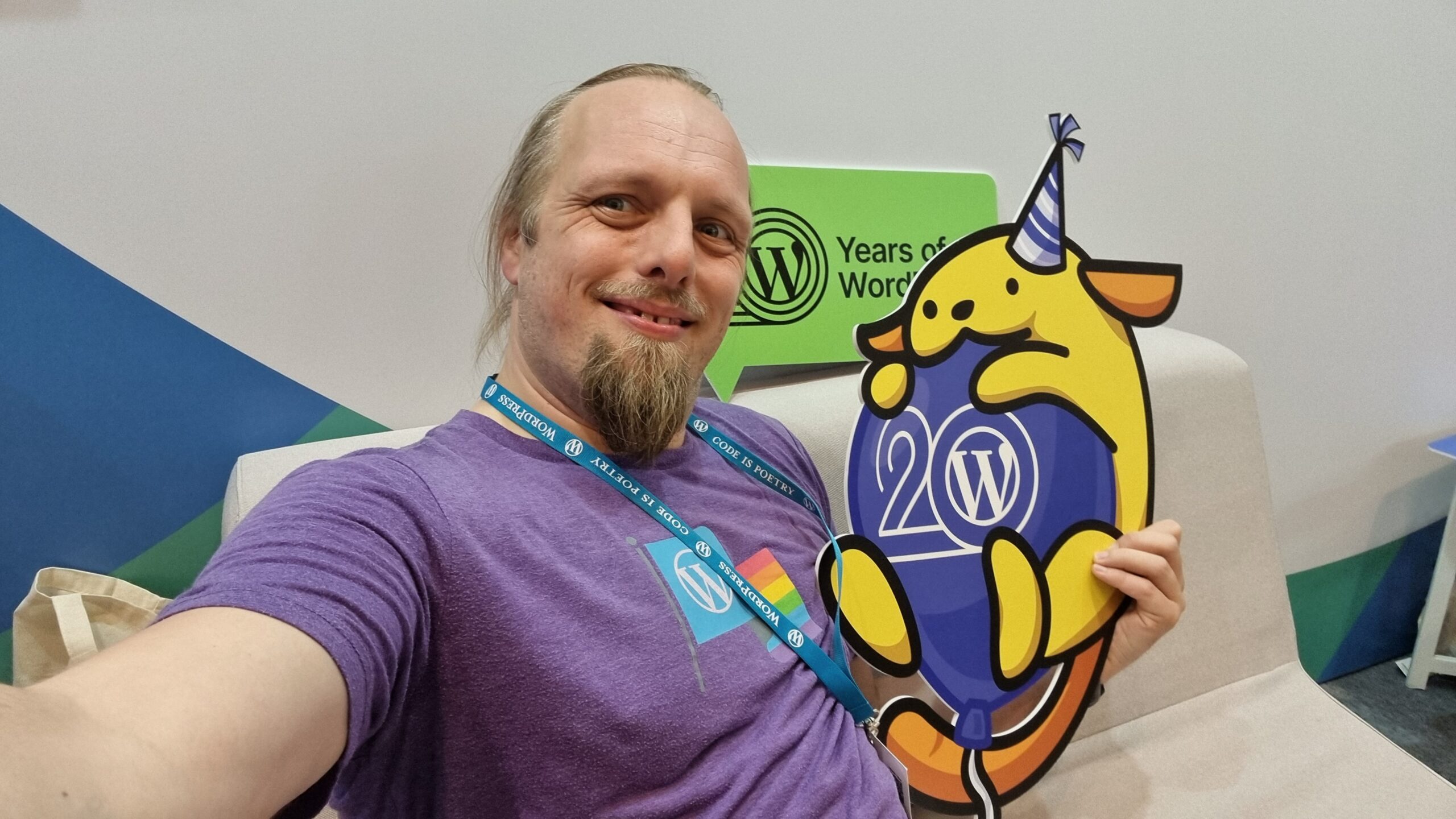Full image of Dan, smiling, wearing a purple t-shirt with a WordPress logo and a Pride flag, hugs a cut-out of a Wappu (itself hugging a "WP 20" balloon and wearing a party hat).
