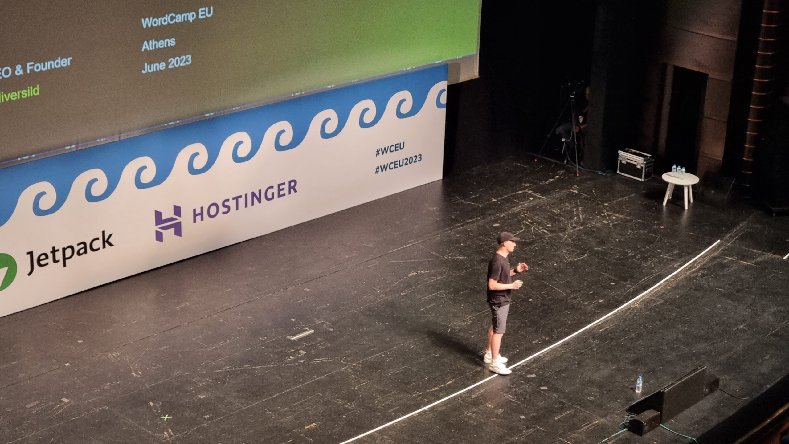 Full image of A man in a literal black hat stands in the centre of a large theatre stage.