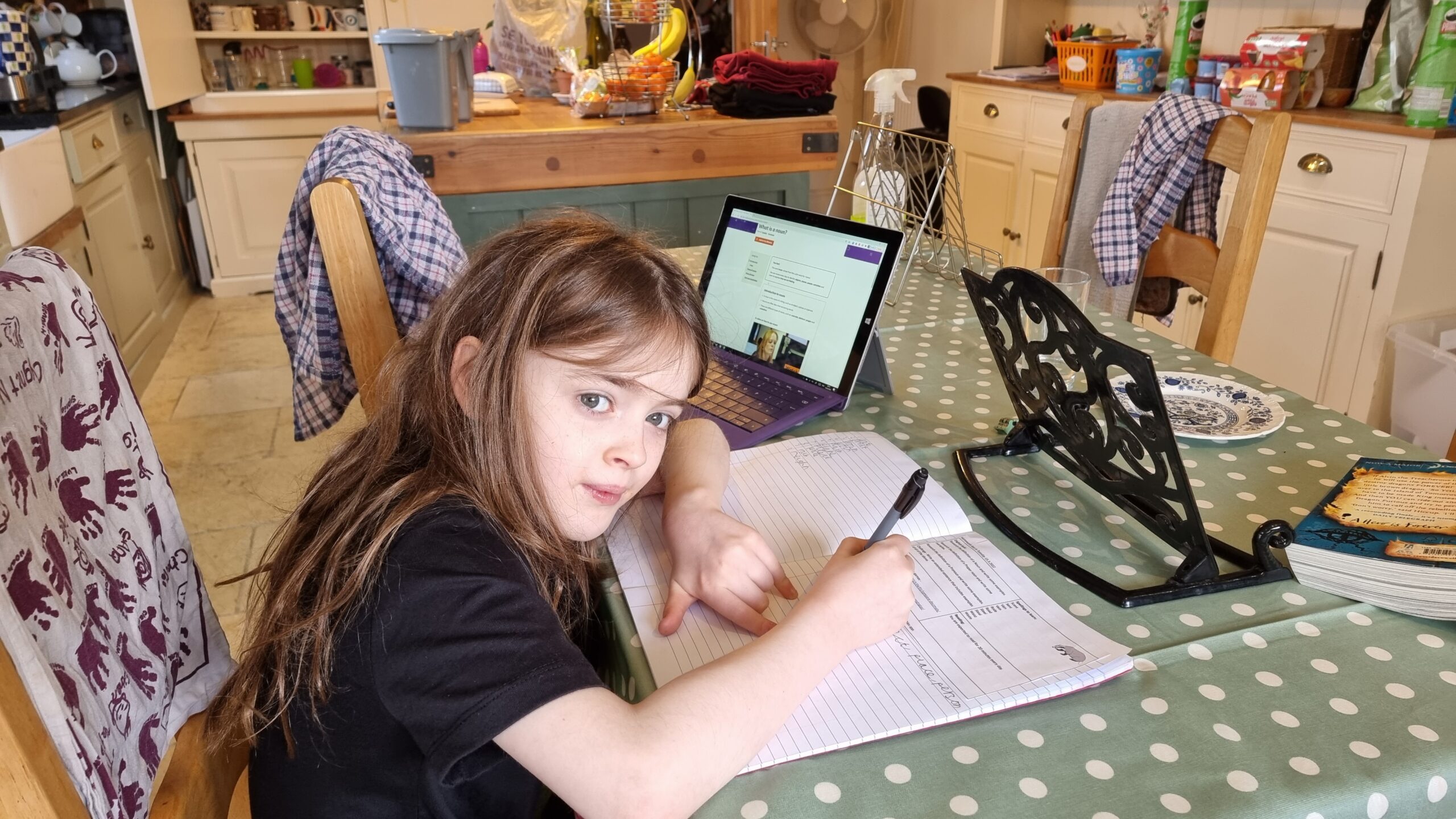Full image of A young girl, her hair wild, sits at a kitchen table with a laptop and a homework book, writing.
