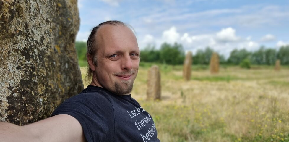 Dan, wearing a black t-shirt with the words "Let's make the web a better place" on, sits with his back to a standing stone. Four more standing stones can be seen stretching away into the bakground, atop a flowery meadow and beneath a slightly cloudy but bright sky.