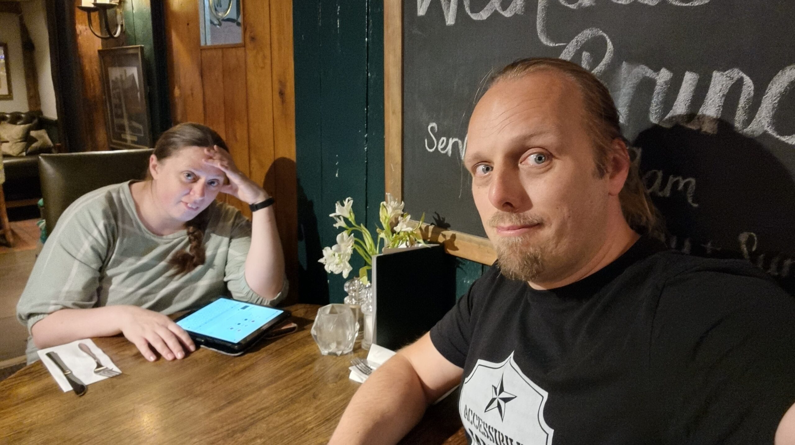 Full image of Ruth and Dan, looking tired and frustrated, sit at a pub table. Ruth is using her tablet computer.