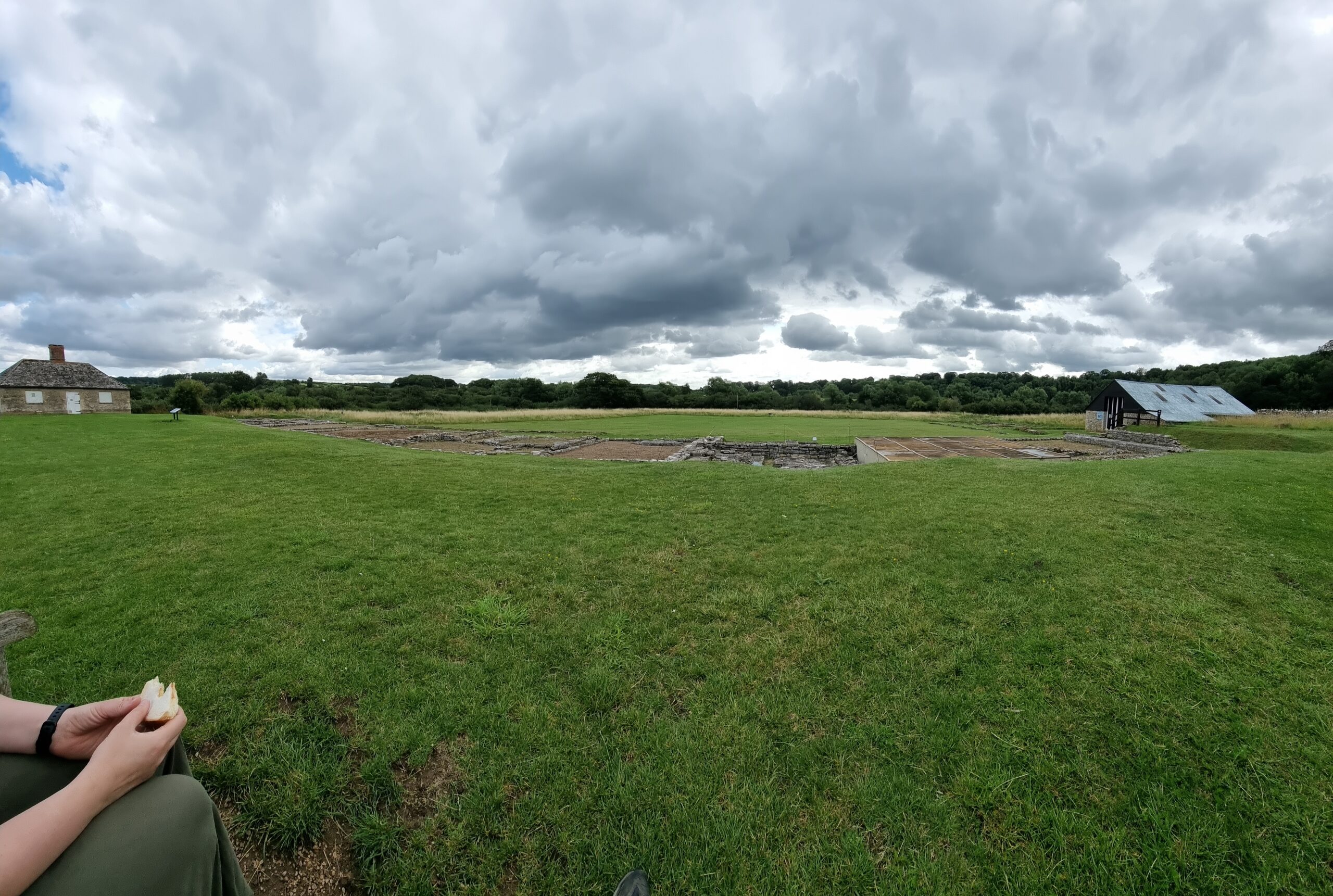 Full image of Panoramic photo showing a field containing the remains of a Roman villa in West Oxfordshire, under grey skies. The walls are barely visible in this wide shot.