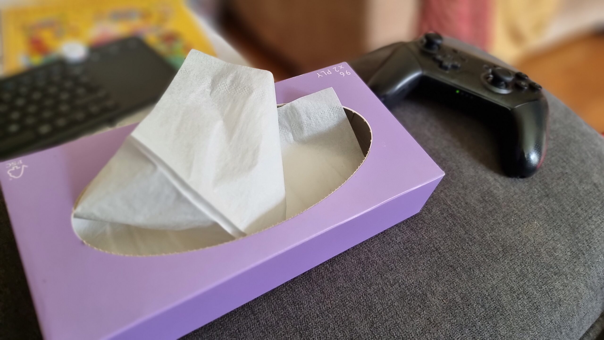 Full image of A box of tissues and a Nintendo Switch Pro Controller on the arm of a sofa.