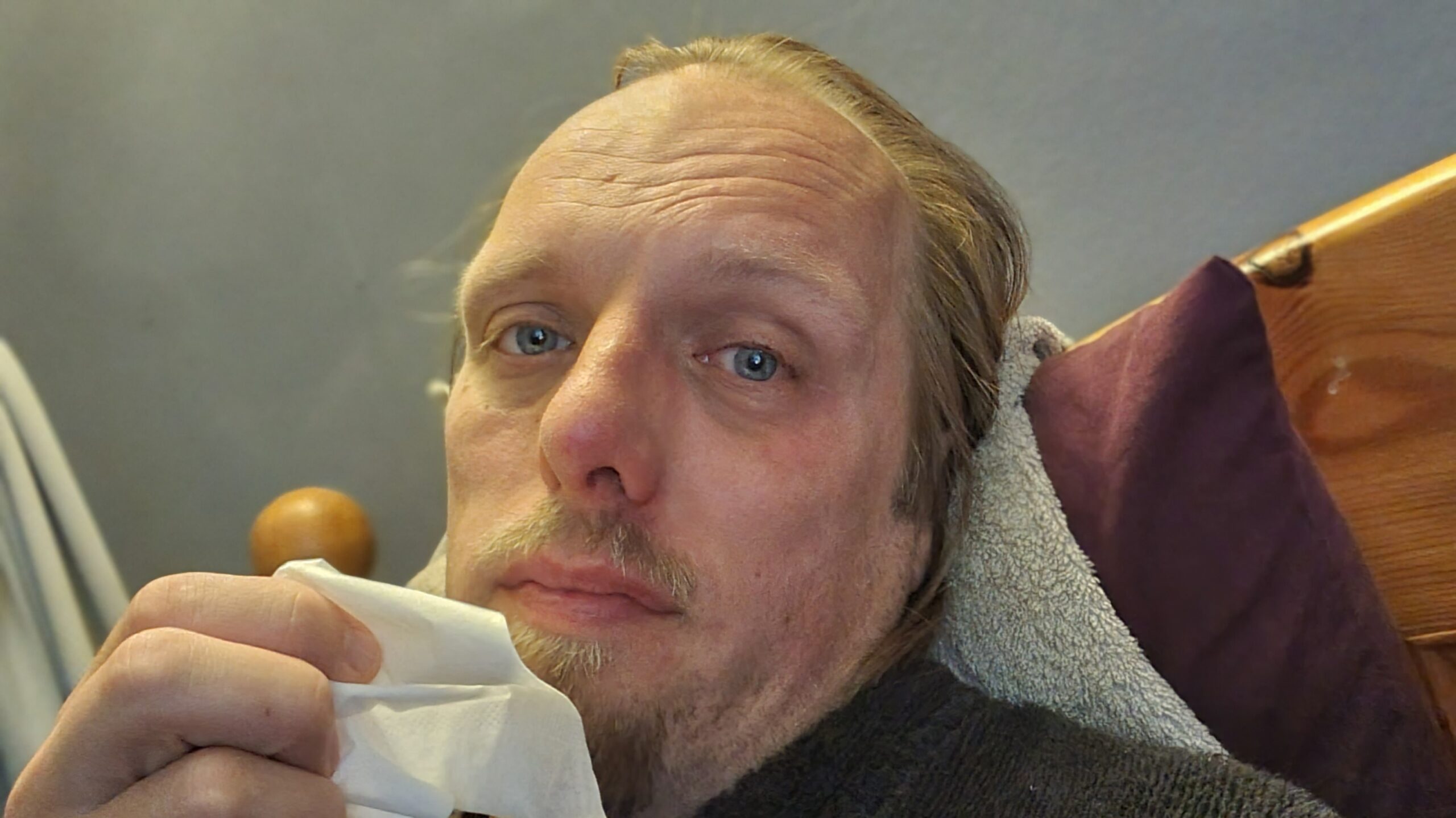 Full image of Dan, sitting in bed, holding a tissue and looking unwell.