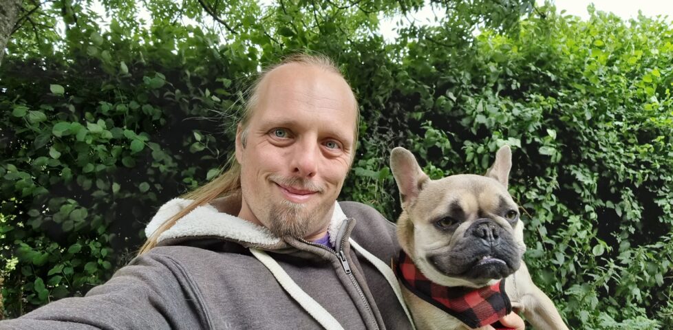 Dan, wearing a grey Three Rings hoodie, carrying French Bulldog Demmy, standing on a path with trees in the background.