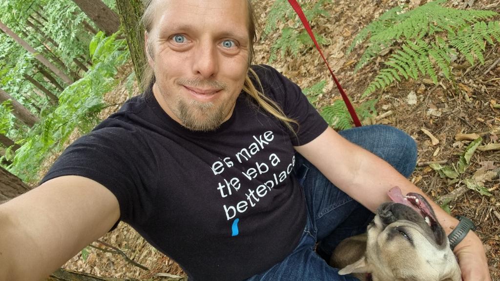 Dan crouches in a forest; a French Bulldog is stretching up to lick his arm.