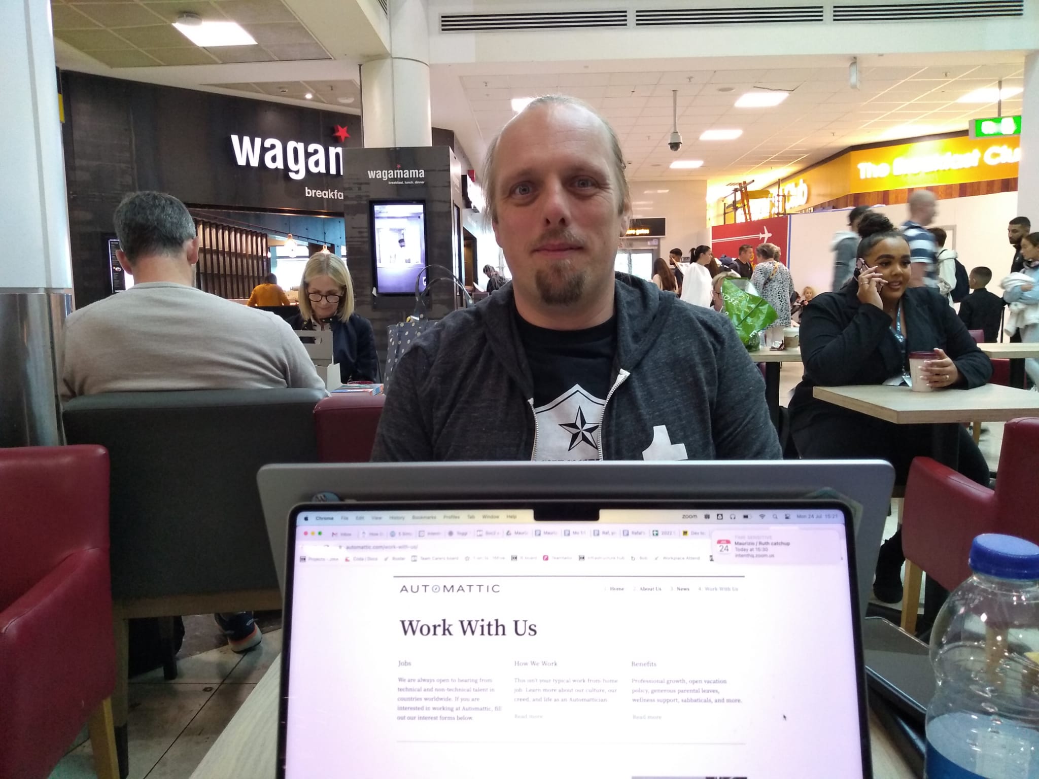 Full image of A laptop screen shows Automattic's "Work With Us" web page. Beyond it, in an airport departure lounge (with diners of Wagamama and The Breakfast Club in the background), Dan sits at another laptop, wearing a black "Accessibility Woke Platoon" t-shirt and grey Tumblr hoodie.