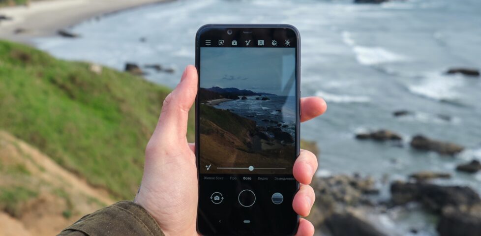 Photo showing a mobile phone, held in a hand, being used to take a photograph of a rugged coastline landscape.