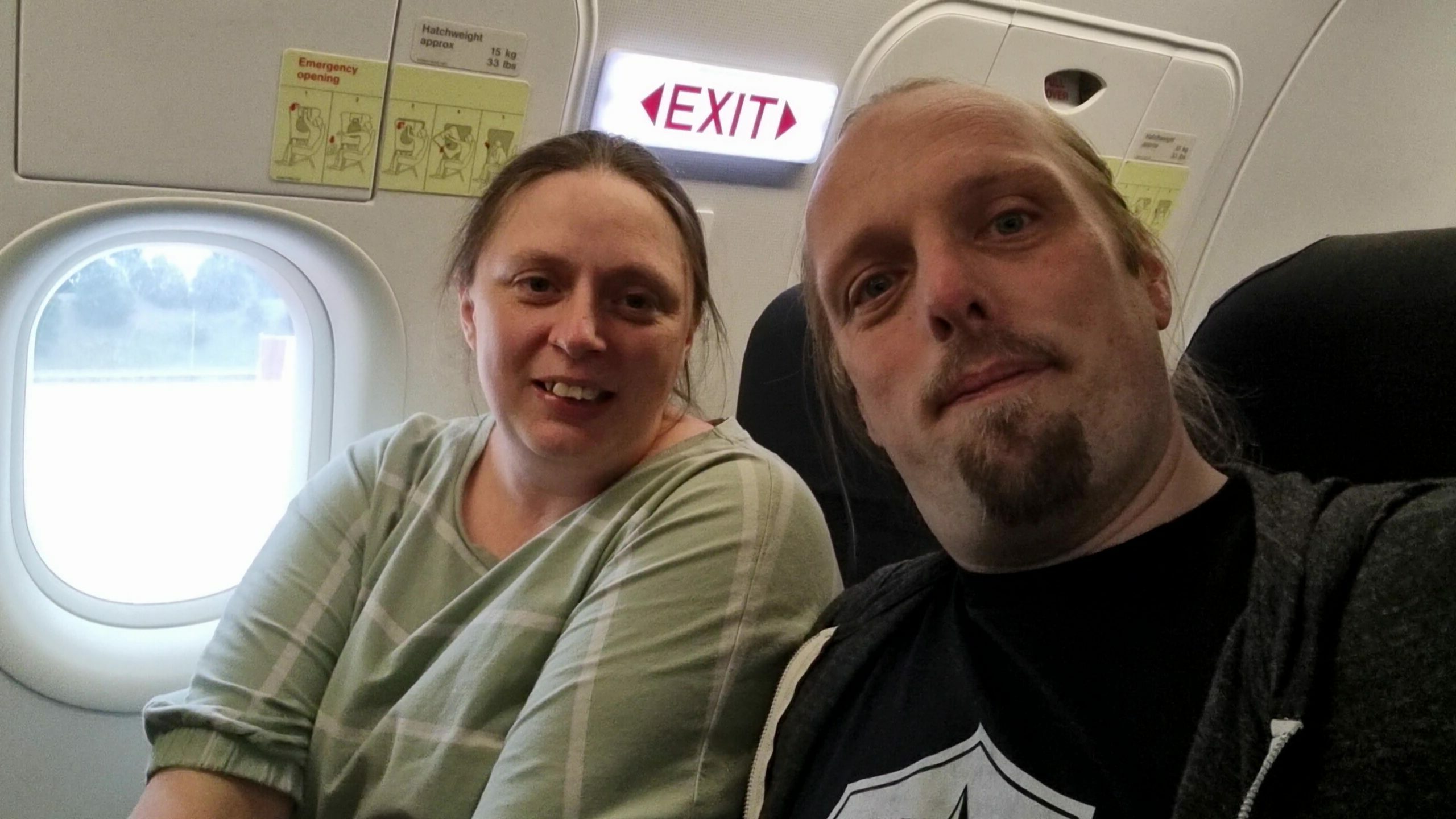 Full image of Ruth, wearing a green top with white stripes, sits alongside Dan, wearing a black t-shirt and grey hoodie, by the wingside emergency exits in an aeroplane.