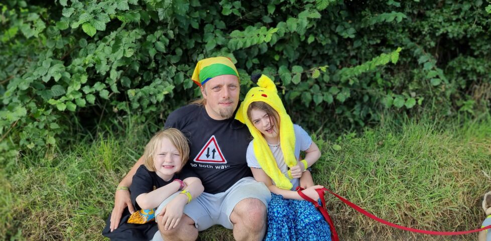 Photograph showing Dan, wearing a "go both ways" t-shirt and a rainbow-striped bandana and grey shorts, sits between a 6-year-old boy and a 9-year-old girl (the girl is wearing a Pikachu hat).