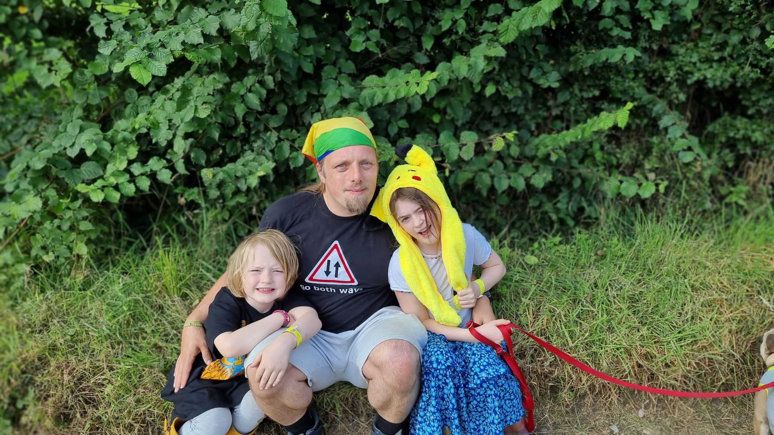 Full image of Photograph showing Dan, wearing a "go both ways" t-shirt and a rainbow-striped bandana and grey shorts, sits between a 6-year-old boy and a 9-year-old girl (the girl is wearing a Pikachu hat).