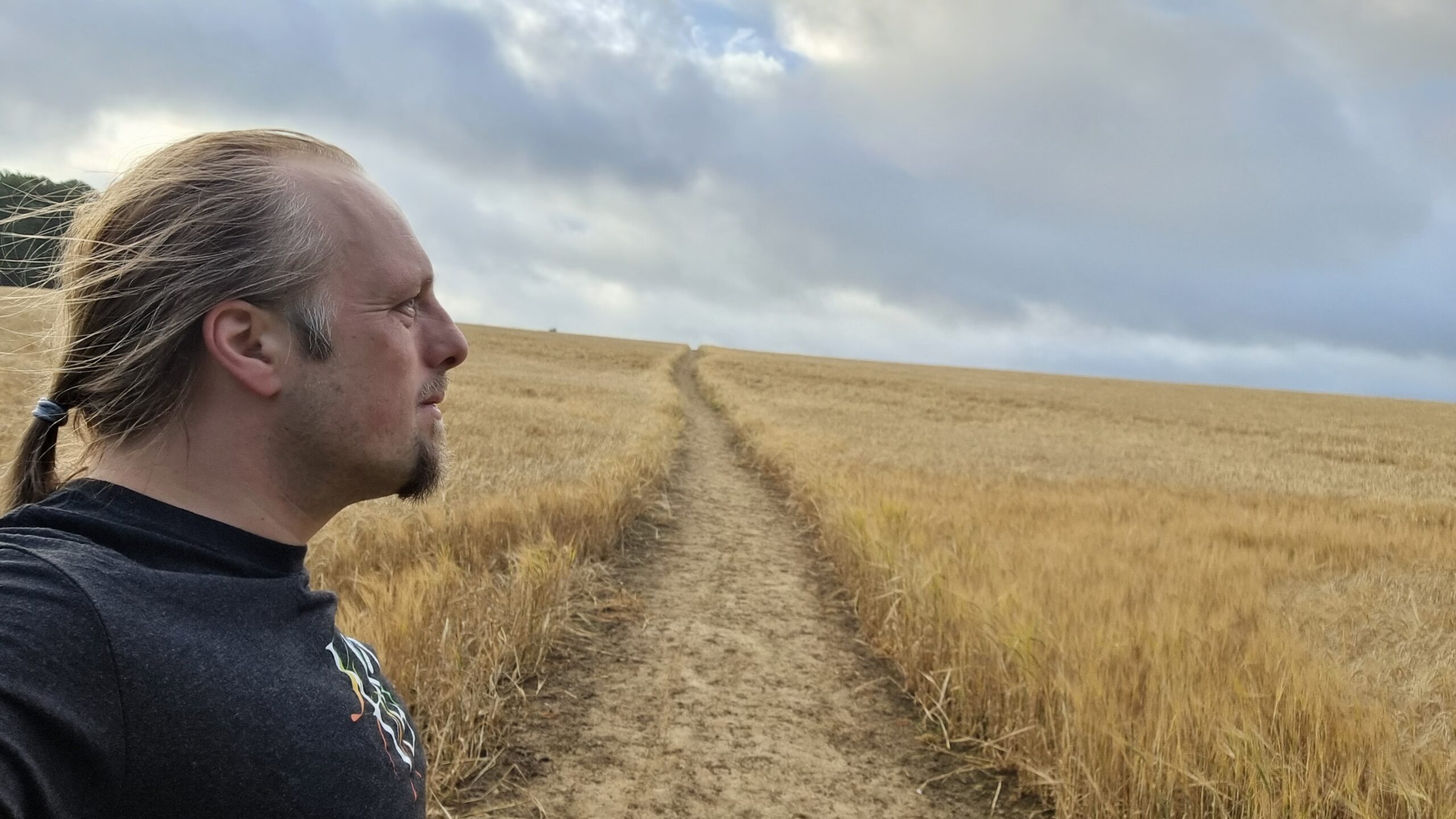 Full image of Dan, on a path through a young cornfield, stares towards the distant clouds.