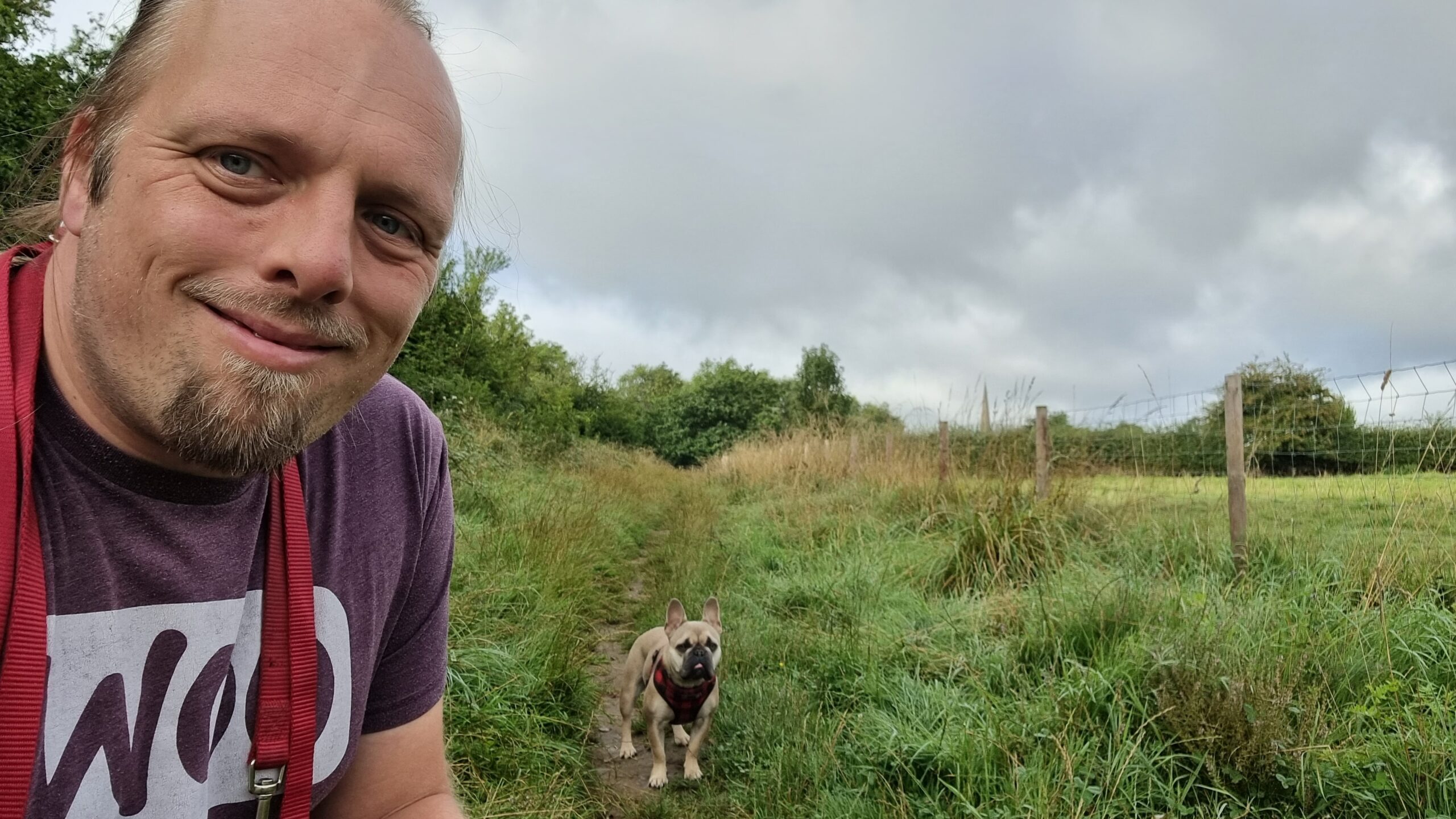 Full image of Dan, wearing a purple "Woo" t-shirt, crouches by Demmy, a champagne-coloured French Bulldog, in a path through the long grass between a wood and a field. In the distance, the church spire of St Peter & St Paul at Church Hanborough can be seen protuding above the treeline.