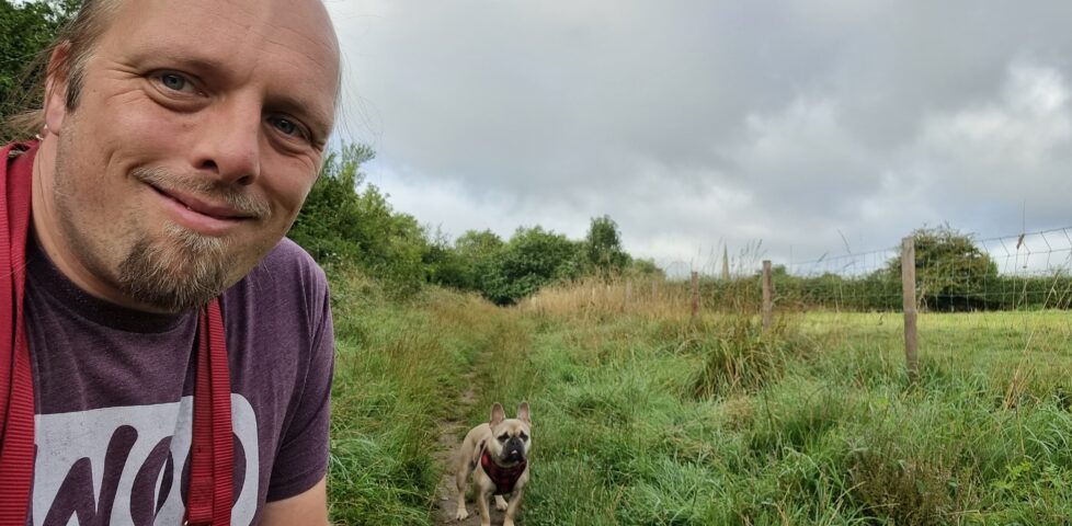 Dan, wearing a purple "Woo" t-shirt, crouches by Demmy, a champagne-coloured French Bulldog, in a path through the long grass between a wood and a field. In the distance, the church spire of St Peter & St Paul at Church Hanborough can be seen protuding above the treeline.