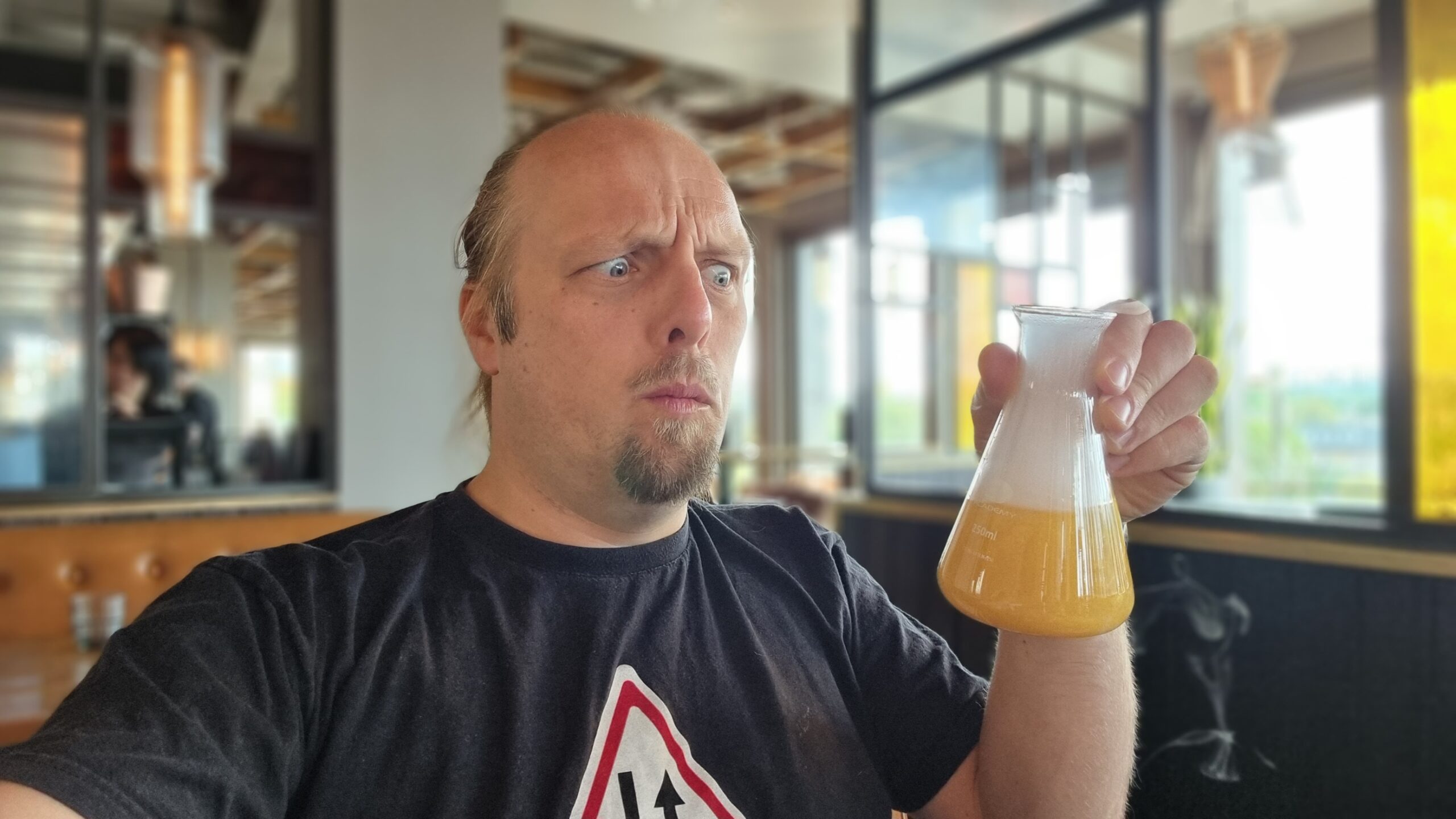 Full image of Selfie photograph of Dan, in a bar with a rooftop view of daylight out the windows in the background, looks concerned as he stares at the a frothy, bubbling flask of yellow liquid he's holding.