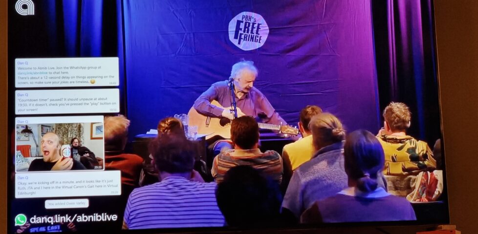 Phootgraph of a wall-mounted television screen. On the screen, comedian Peter Buckley Hill sits with his guitar on his lap in front of an audience: the "PBH's Free Fringe" logo is on the curtain behind him. On the left of the screen a series of WhatsApp messages appear, including one showing a photo of Dan holding a can of Old Speckled Hen beer.