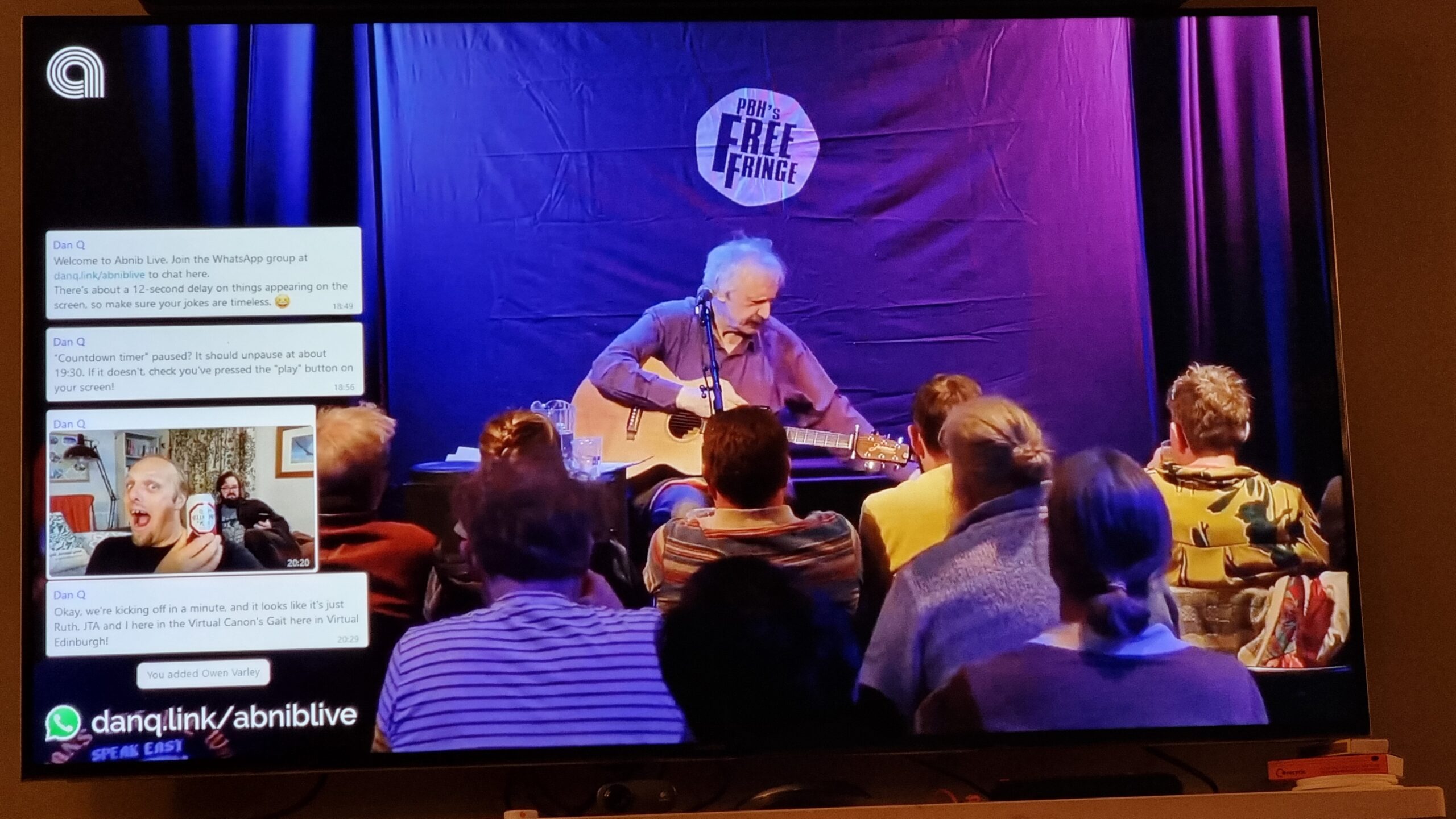Full image of Phootgraph of a wall-mounted television screen. On the screen, comedian Peter Buckley Hill sits with his guitar on his lap in front of an audience: the "PBH's Free Fringe" logo is on the curtain behind him. On the left of the screen a series of WhatsApp messages appear, including one showing a photo of Dan holding a can of Old Speckled Hen beer.