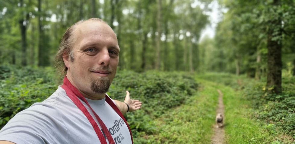 Dan, wearing a white t-shirt and with a red dog lead hanging over his shoulder, stands in a forest, gesturing down a path to a small French Bulldog following him.