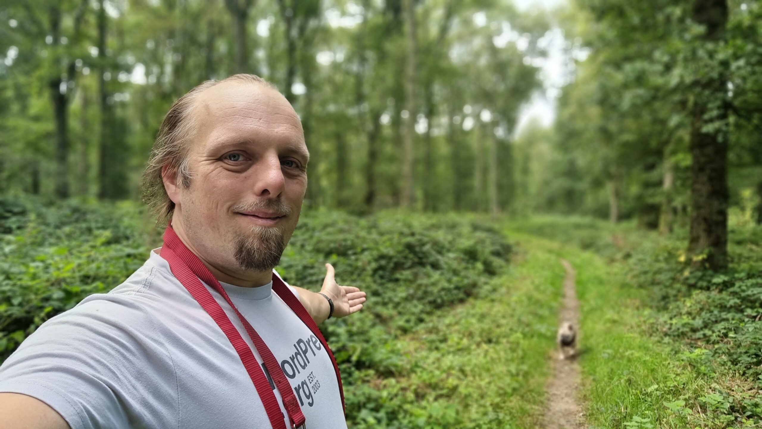 Full image of Dan, wearing a white t-shirt and with a red dog lead hanging over his shoulder, stands in a forest, gesturing down a path to a small French Bulldog following him.