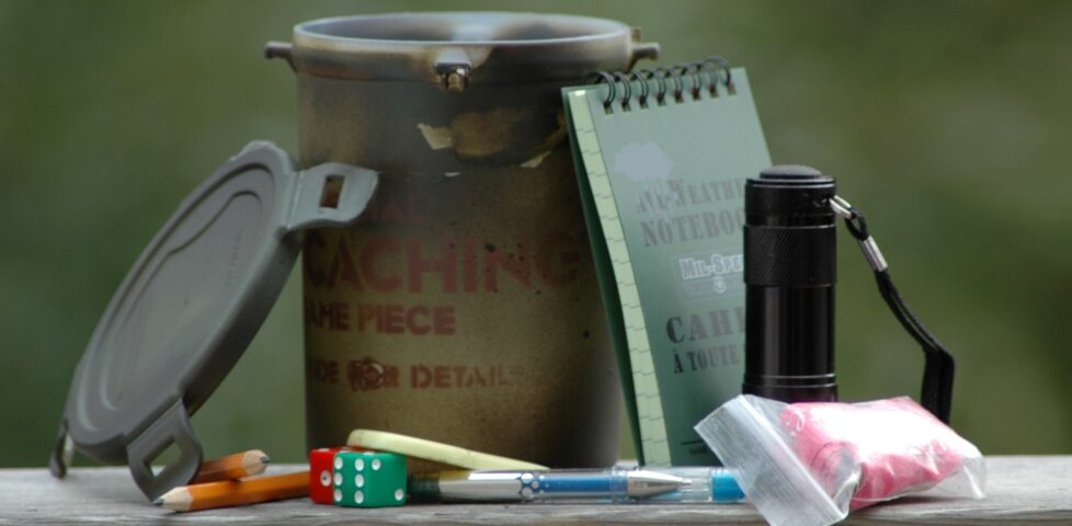 Photograph showing a medium-sized geocache container with its contents laid-out around it: various pieces of swag for trade, plus a notebook.