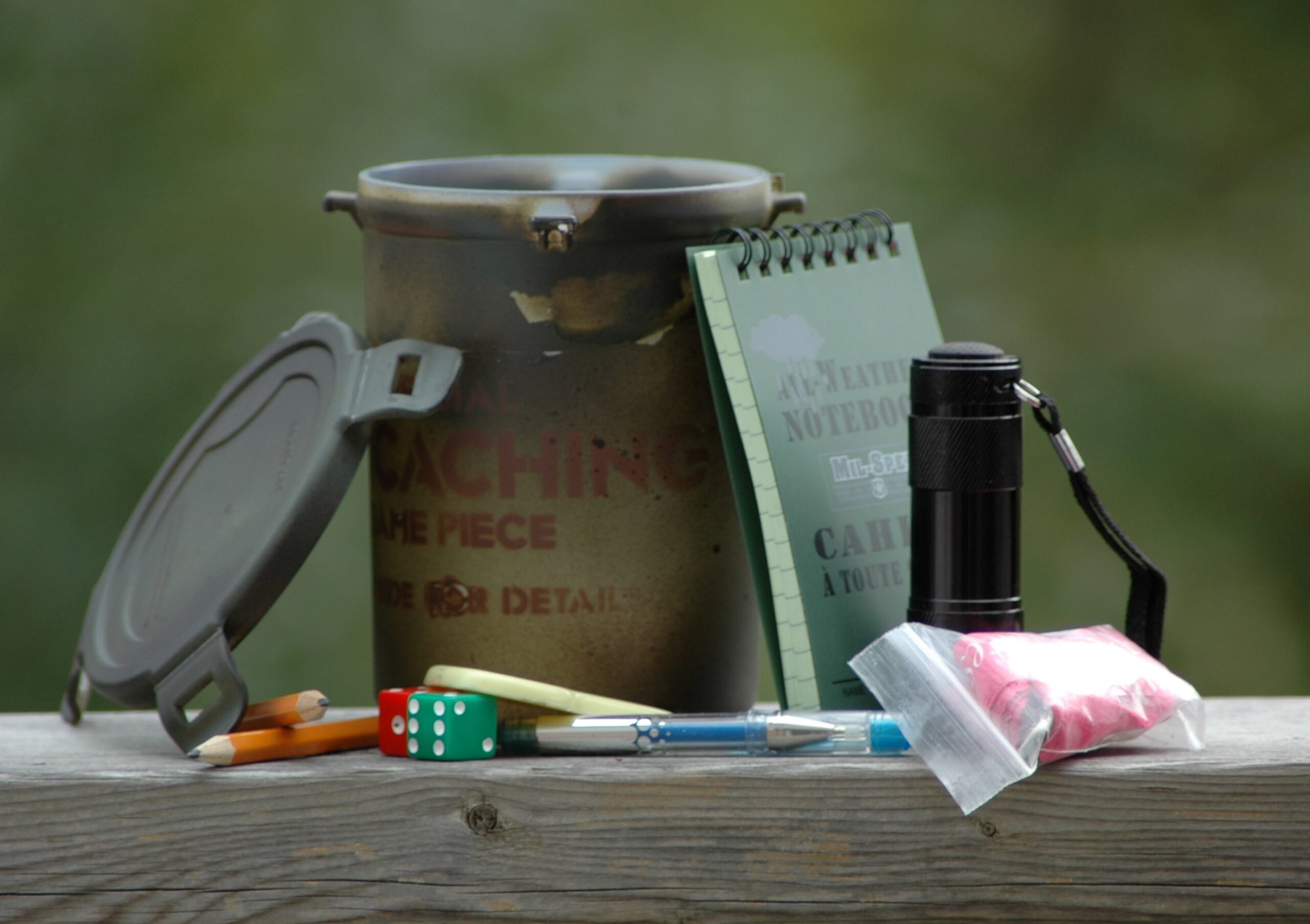 Full image of Photograph showing a medium-sized geocache container with its contents laid-out around it: various pieces of swag for trade, plus a notebook.