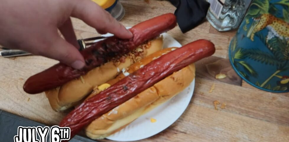 Frame from a video showing TomSka's hand pushing a hot dog sausage into a bun, on top of fried onions and sauces. The picture is captioned "July 6th".