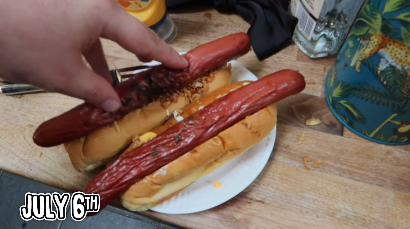Full image of Frame from a video showing TomSka's hand pushing a hot dog sausage into a bun, on top of fried onions and sauces. The picture is captioned "July 6th".