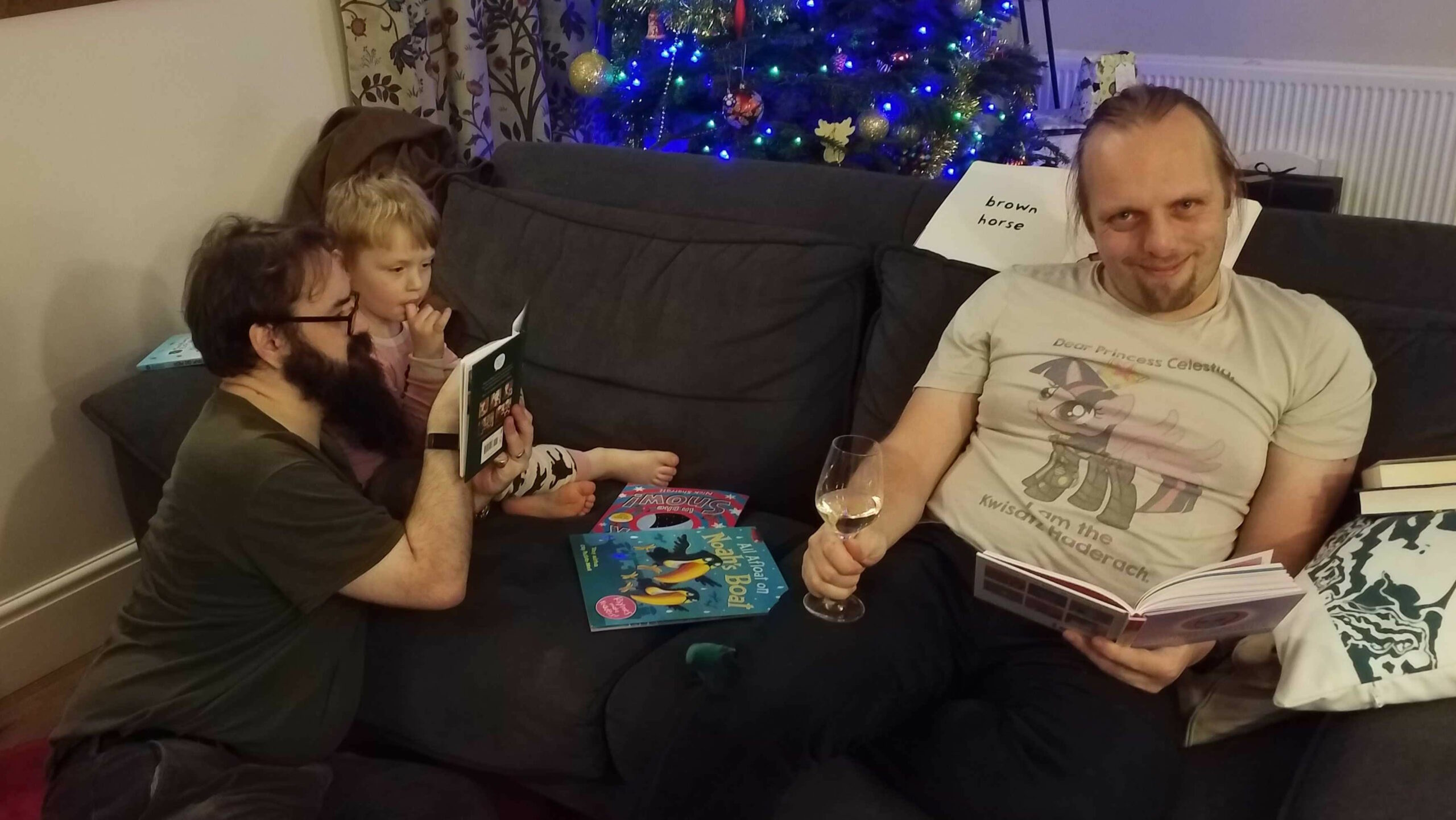 Full image of Dan, wearing a Princess Twilight Sparkle / Frank Herbert's Dune crossover fan art t-shirt, sits on a grey sofa in front of a lit Christmas tree, holding a glass of wine. At the other end of the sofa JTA, a white man with a thick beard and glasses, reads to a (tired-looking) young boy. All three are surrounded by books.