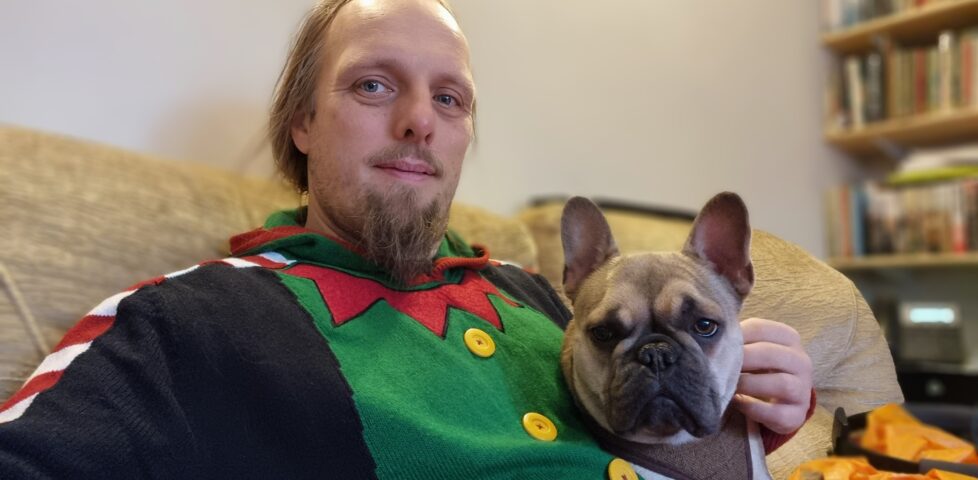 Dan, wearing an "elf costume" Christmas jumper, looks into the camera while cuddling a French Bulldog. The pair are sitting on a beige sofa.