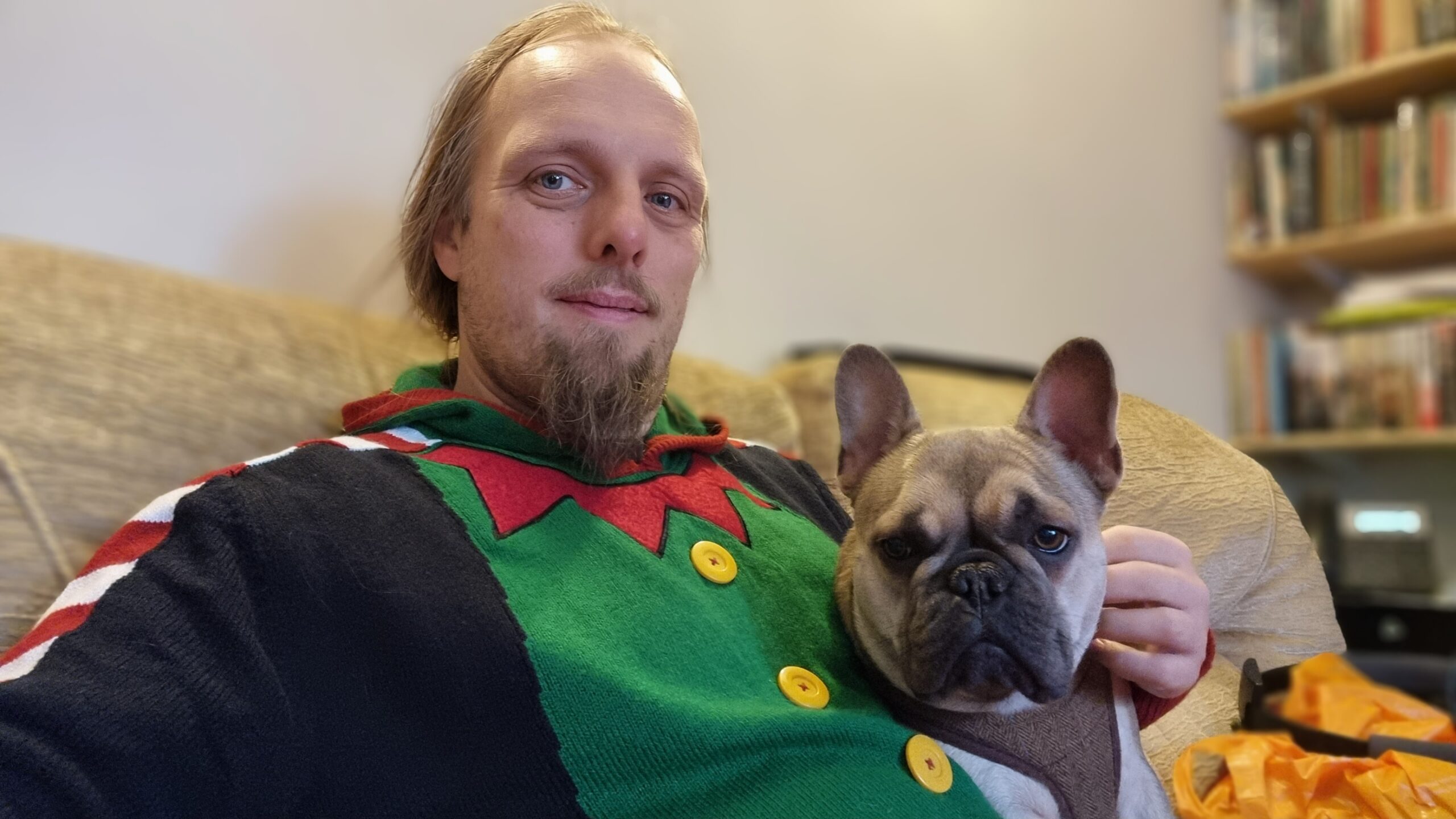 Full image of Dan, wearing an "elf costume" Christmas jumper, looks into the camera while cuddling a French Bulldog. The pair are sitting on a beige sofa.