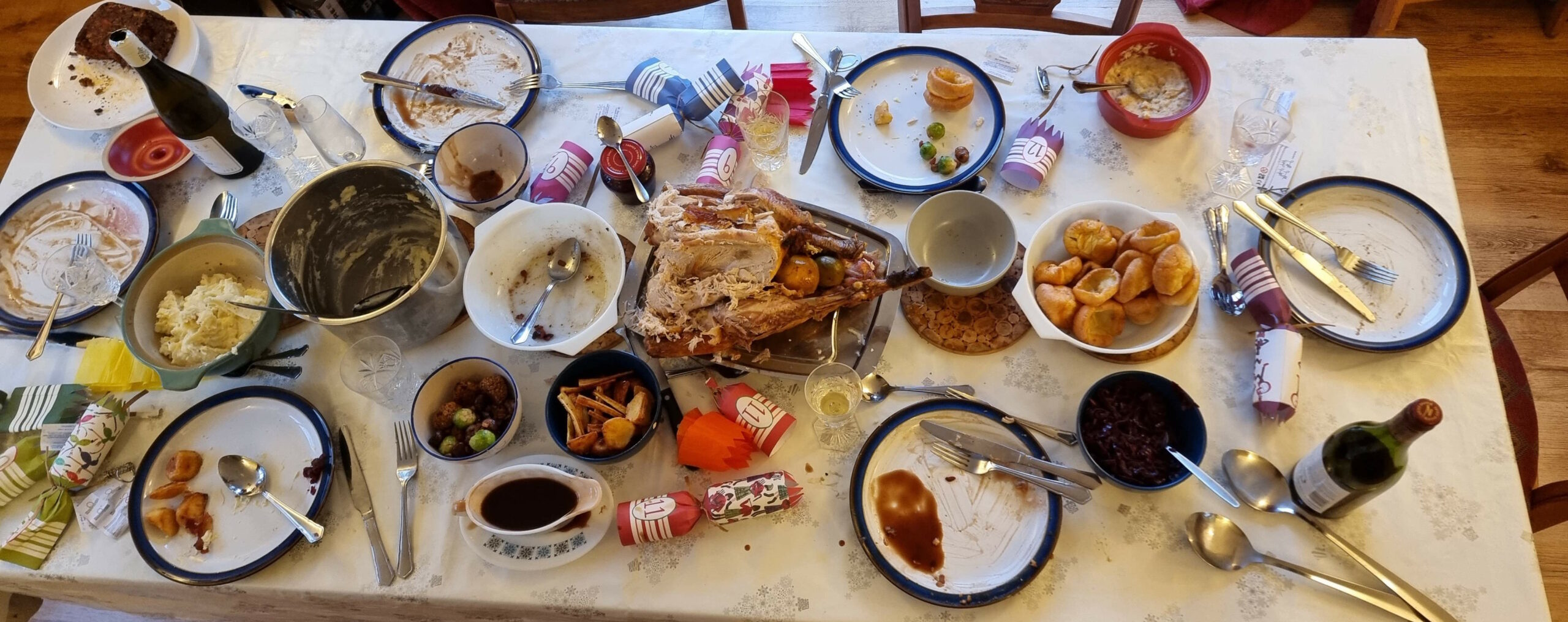 Full image of Top-down view of a dining table set with a Christmas-themed tablecloth. The meal has concluded and the seats have been vacated, but large amounts of food - most of a turkey, half a nutloaf, lots of mashed potato, several sprouts, stuffing balls, and chestnuts, some roast potatoes and parsnips, an entire boat full of gravy, and almost a dozen Yorkshire puddings - are still set out.