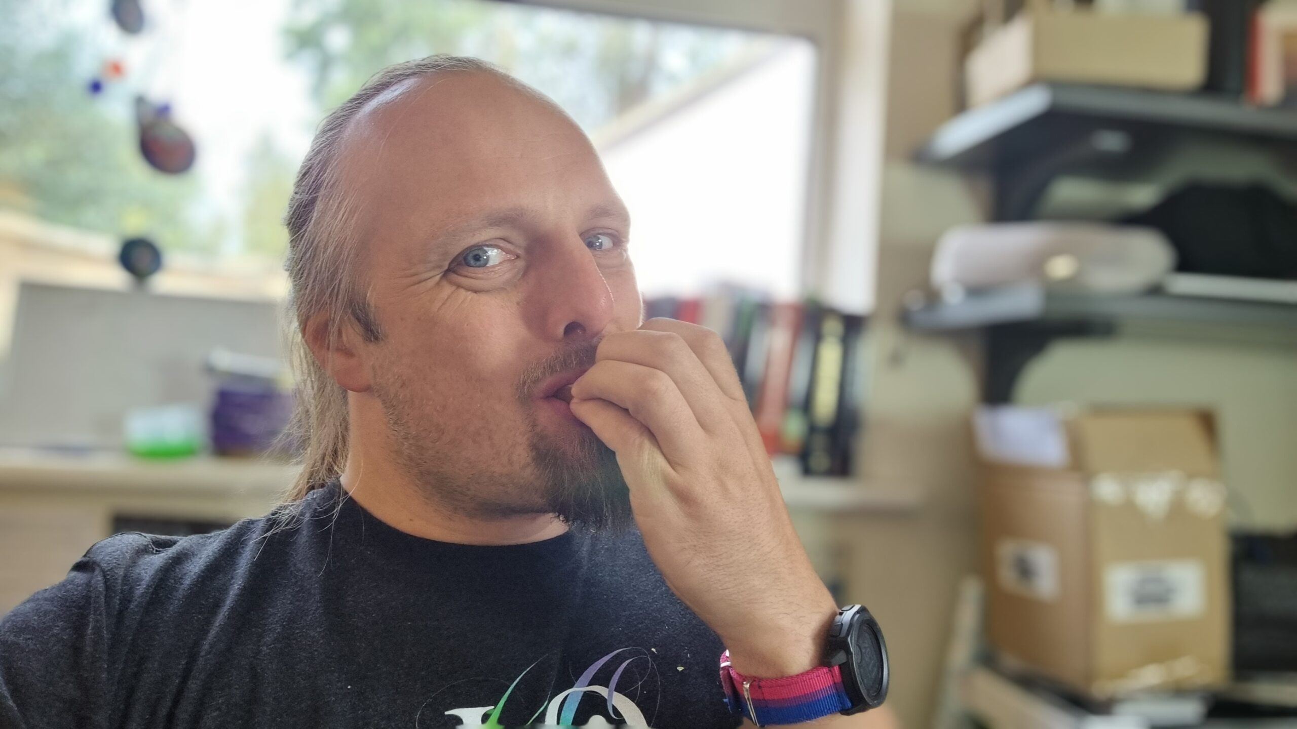Full image of Dan - a white man with long hair tied back and a goatee-style beard - bites into something concealed by his hand (but implied to be a crisp sandwich), which is at his mouth. He's wearing a black t-shirt on which the top part of a swirling rainbow can be made out, and a watch with a "bi pride"-flag coloured watch strap. A window and a cluttered office space are out-of-focus behind him.