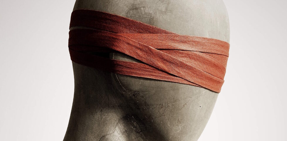 Silhouette photograph of the head of a plain wooden mannequin, wearing a blindfold improvised from a strap of red cloth wrapped repeatedly around its head. Adapted from a photograph by David Underland.
