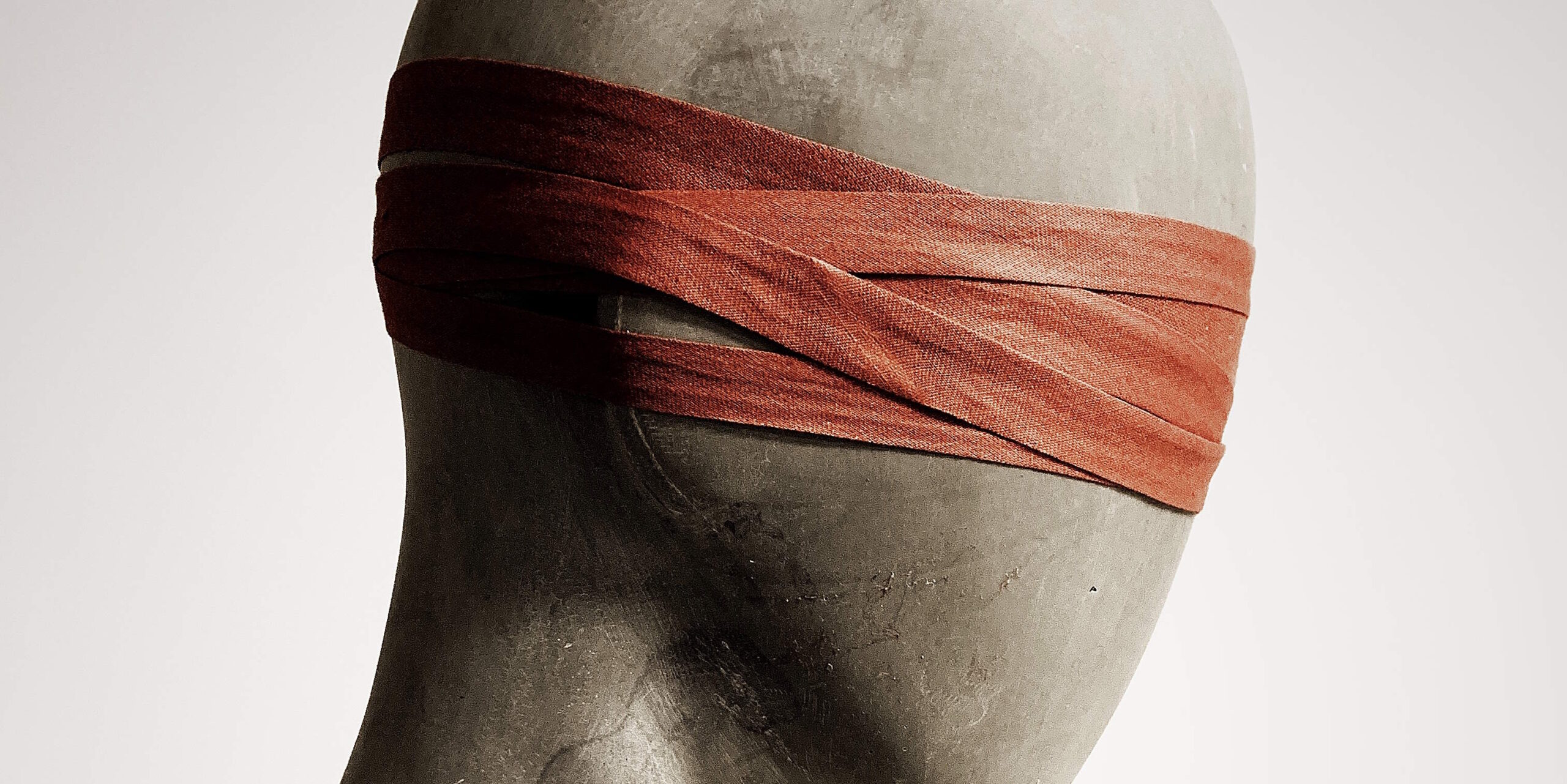 Full image of Silhouette photograph of the head of a plain wooden mannequin, wearing a blindfold improvised from a strap of red cloth wrapped repeatedly around its head. Adapted from a photograph by David Underland.