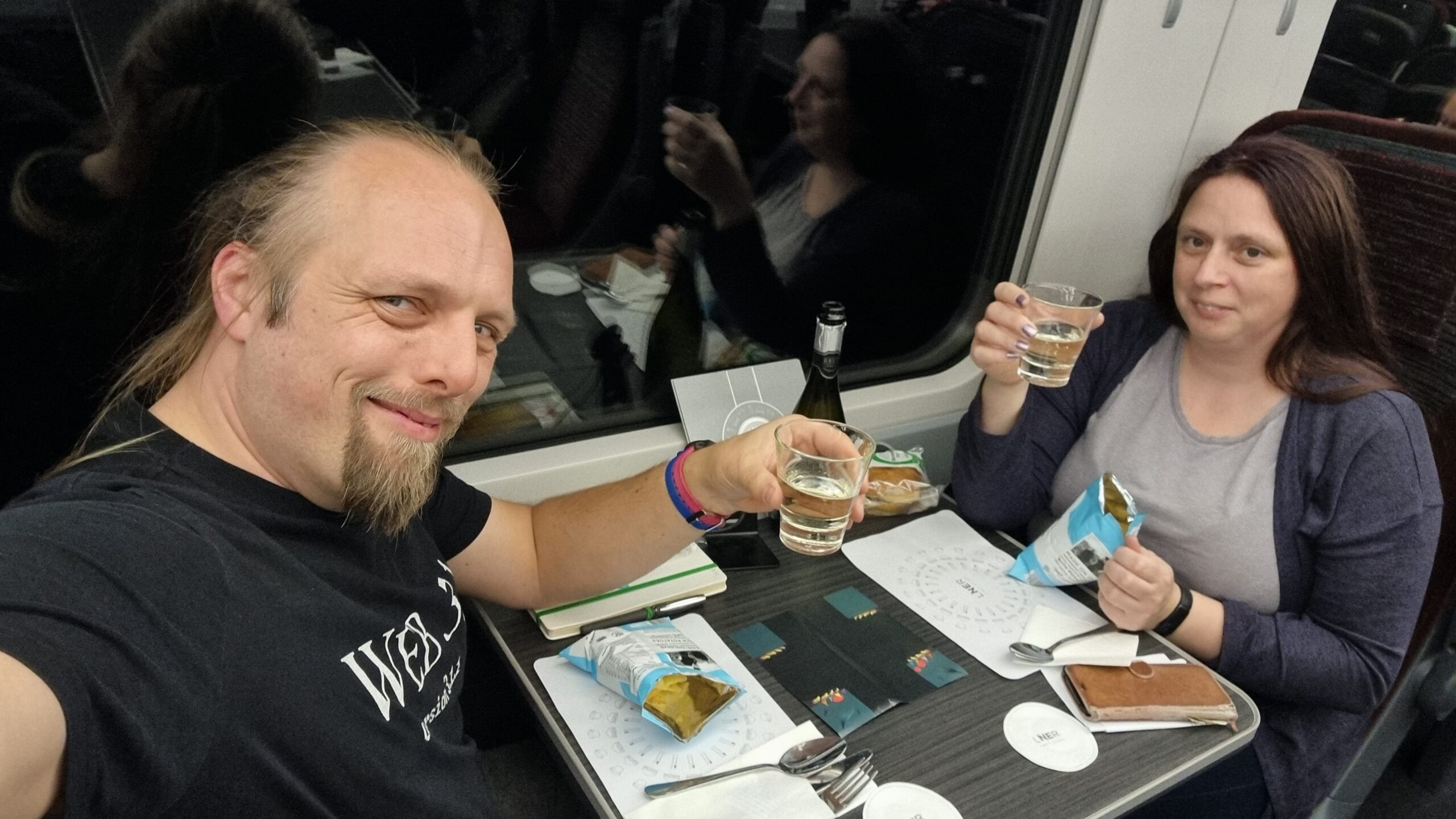Full image of Dan and Ruth sat on opposite sides of a table on a train, with darkness outside the window behind, raising tumbler glasses full of prosecco and smiling.