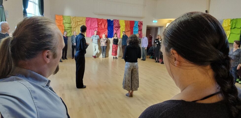 In a church hall, its walls decorated with colourful cloths, Dan and Ruth stand in a large circle of people, watching a man and a woman preparing to demonstrate some tango steps.