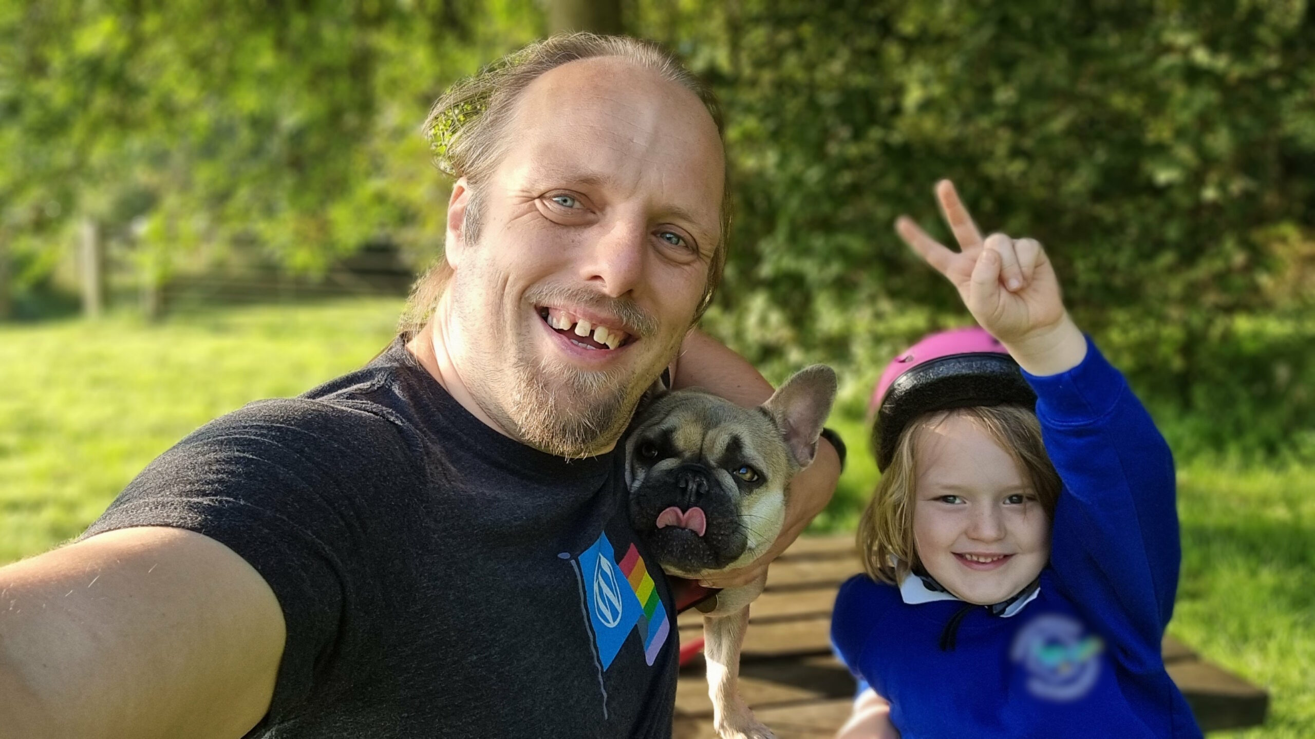 Full image of Dan, wearing a WordPress Pride "rainbow flag on black" t-shirt, sits on a park bench alongside a French Bulldog (with her tongue sticking out) and a young boy (throwing a peace sign, wearing a pink cycle helmet and a blue school uniform).