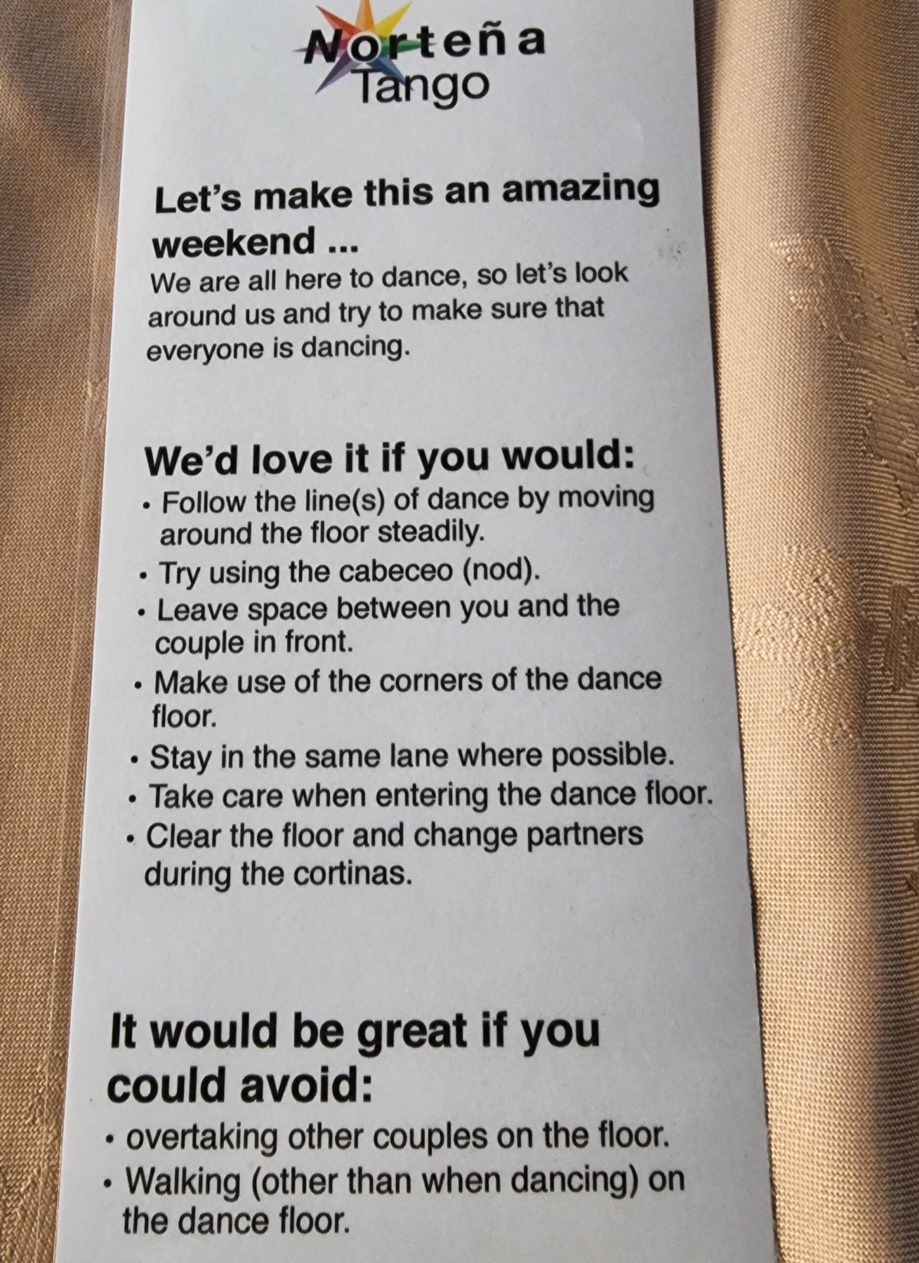 Full image of Photograph of a small laminated instruction sheet on a golden tablecloth. Titled "Norteña Tango", it reads: Let's make this an amazing weekend. We are all here to dance, so let's look around us and try to make sure that everyone is dancing. We'd love it if you would follow the lines of dance by moving around the floor steadily, try using the cabeceo, leave space between you and the couple in front, make use of the corners of the dance floor, stay in the same lane where possible, take care when entering the dance floor, clear the floor and change partners during the cortinas. It would be great if you could avoid overtaking other couples on the floor, walking (other than when dancing) on the floor.