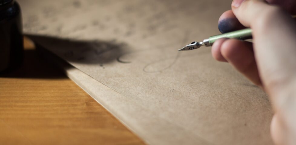 Traditional inkwell and pen, the latter held in an inkstained finger grip, being used to write a letter on unbleached paper atop a wooden desk.