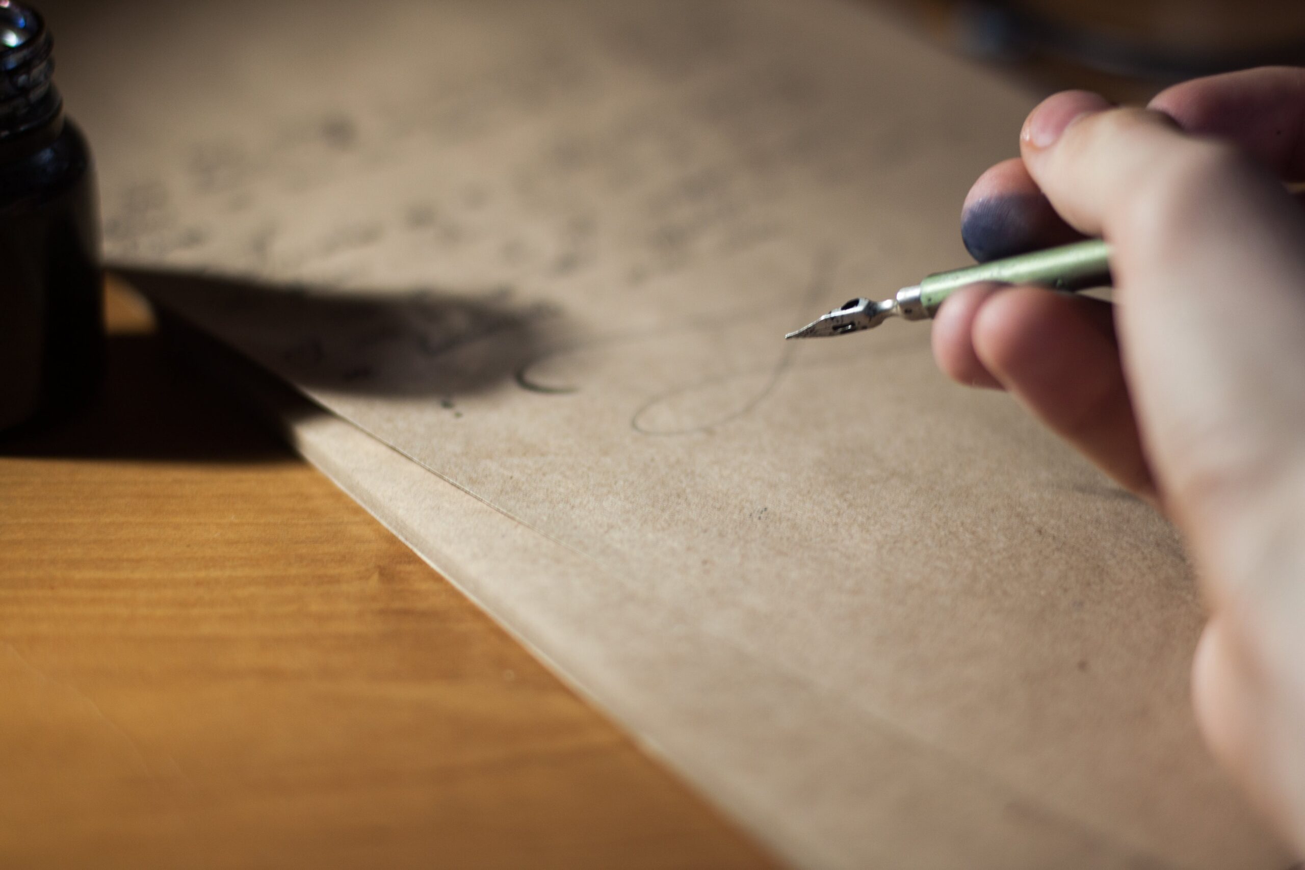 Full image of Traditional inkwell and pen, the latter held in an inkstained finger grip, being used to write a letter on unbleached paper atop a wooden desk.