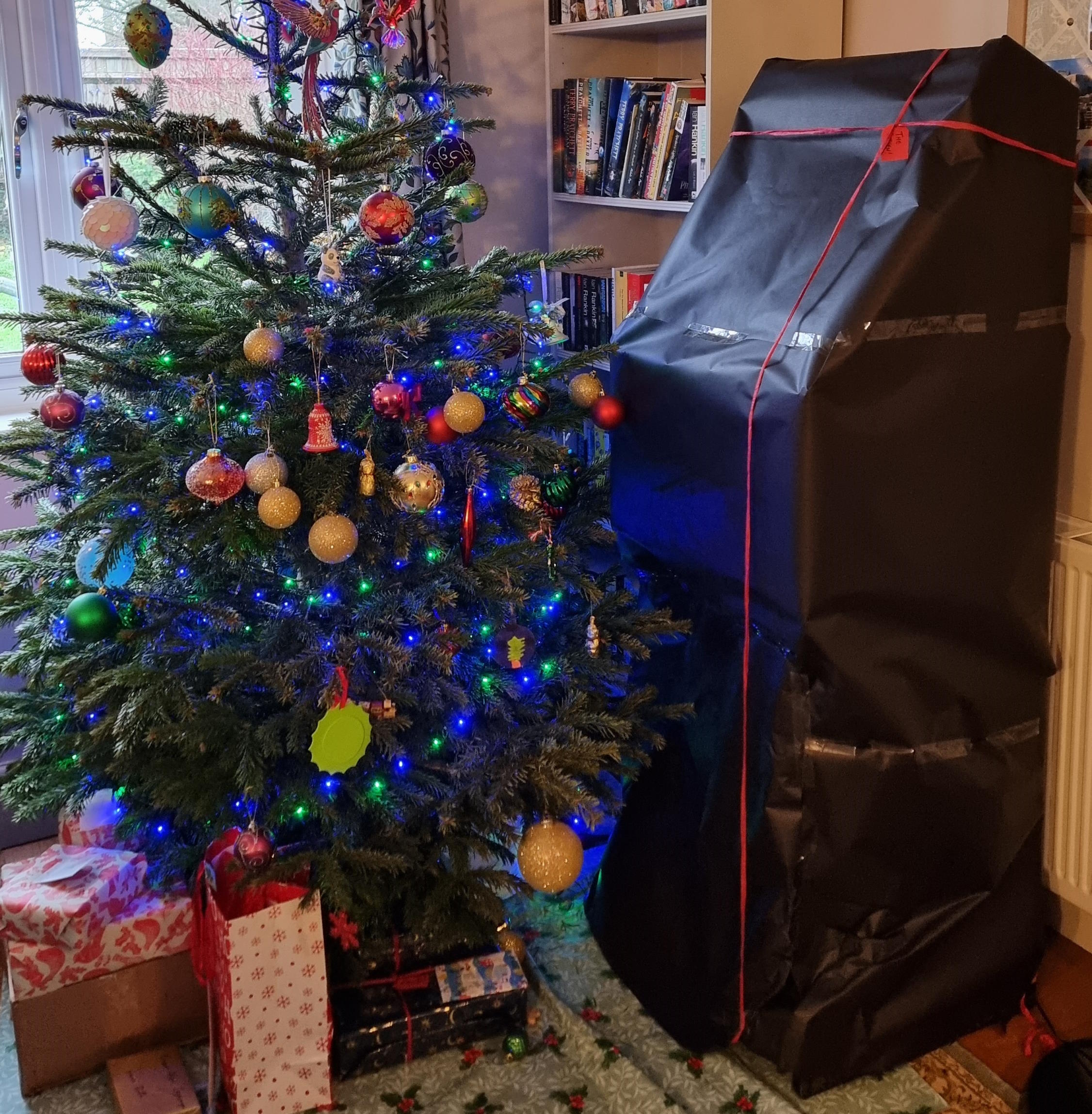 Full image of A large, arcade-cabinet-shaped present, wrapped in black paper and a red ribbon, stands alongside a Christmas tree.