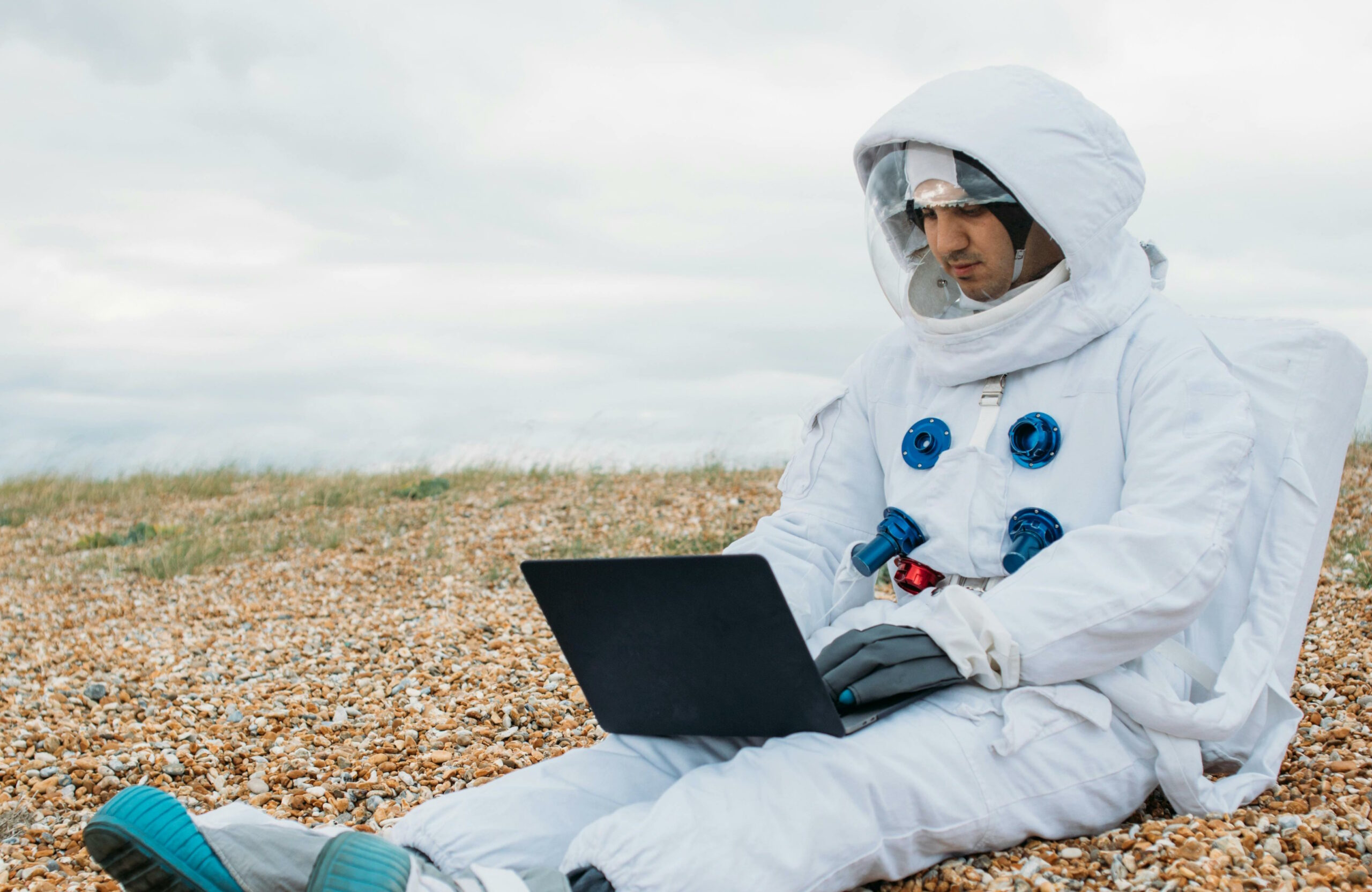 Full image of A white man wearing a spacesuit sits on a pebble beach using a laptop.