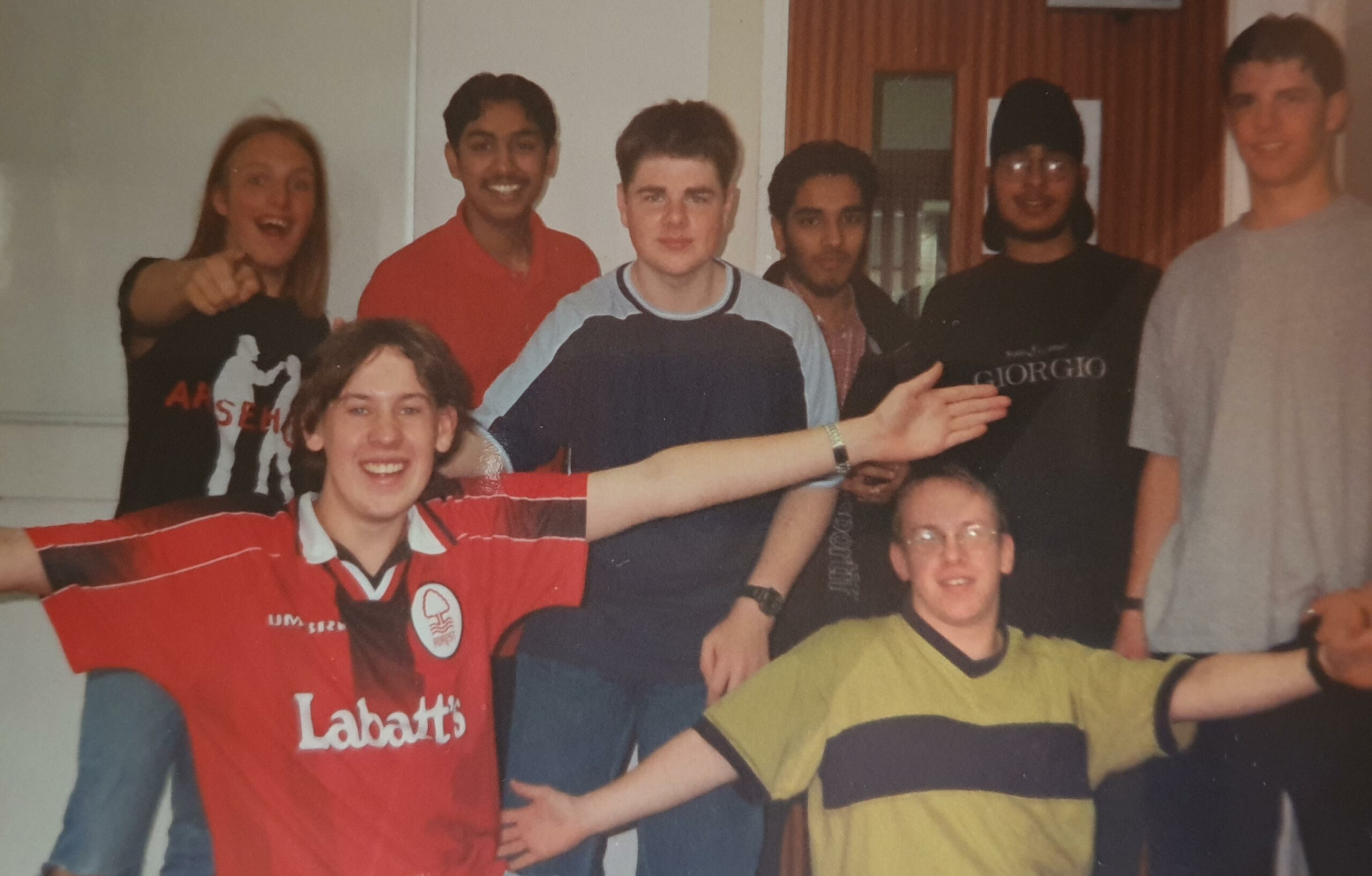 Full image of A young Dan, plus seven other casually-dressed young men, pose in a classroom.