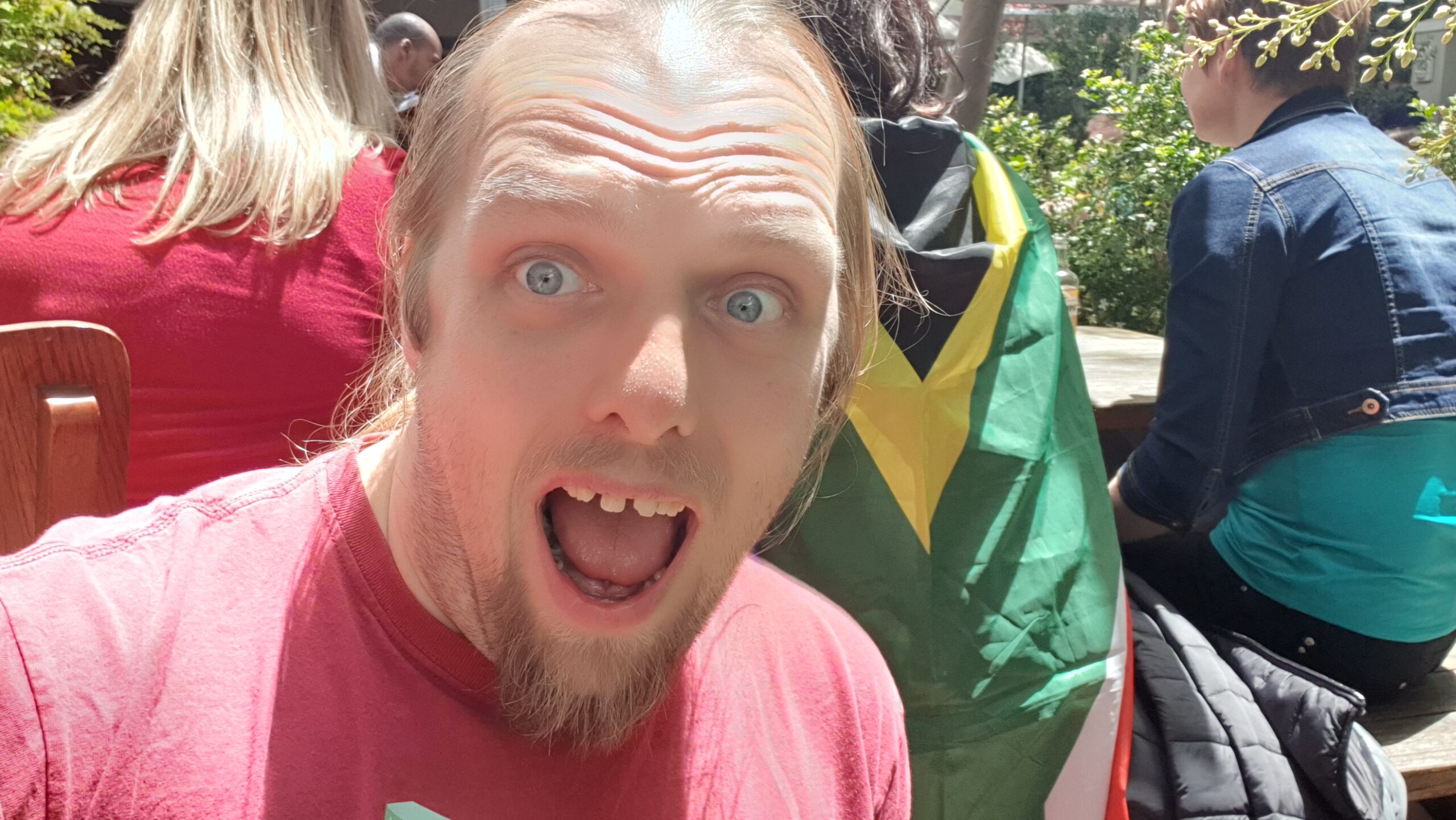 Full image of Dan, wearing a red t-shirt and in bright sunshine, pretends (badly) to be shouting excitedly. In the background, a group of people are watching a televised rugby match; one is wearing a South African flag as a cape.