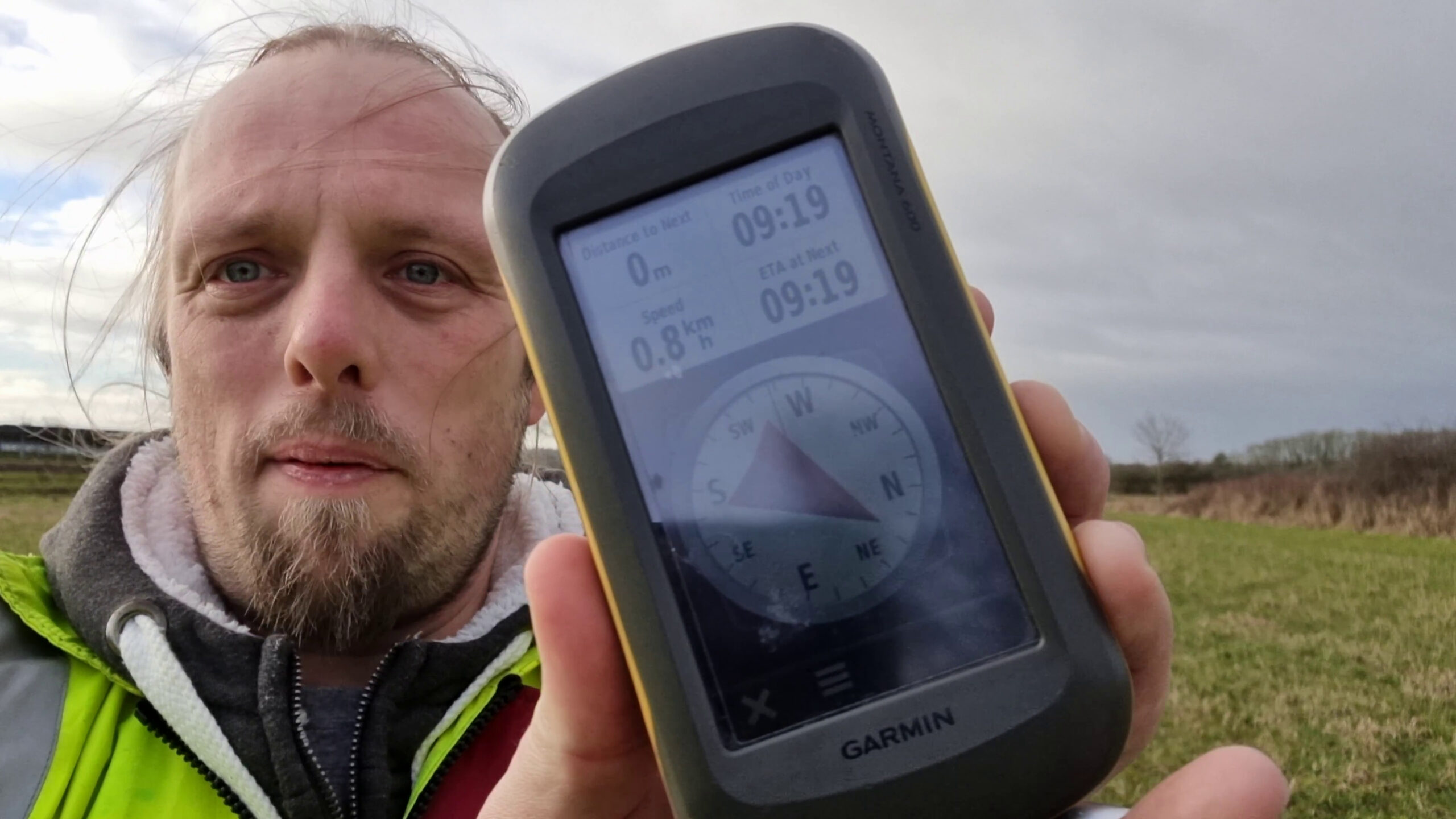 Full image of Dan, outdoors in a field on a grey day and with the wind whipping his hair across his face, wearing a high-vis jacket over a warm fleece, holds up a GPS receiver which shows he's zero metres from his destination.